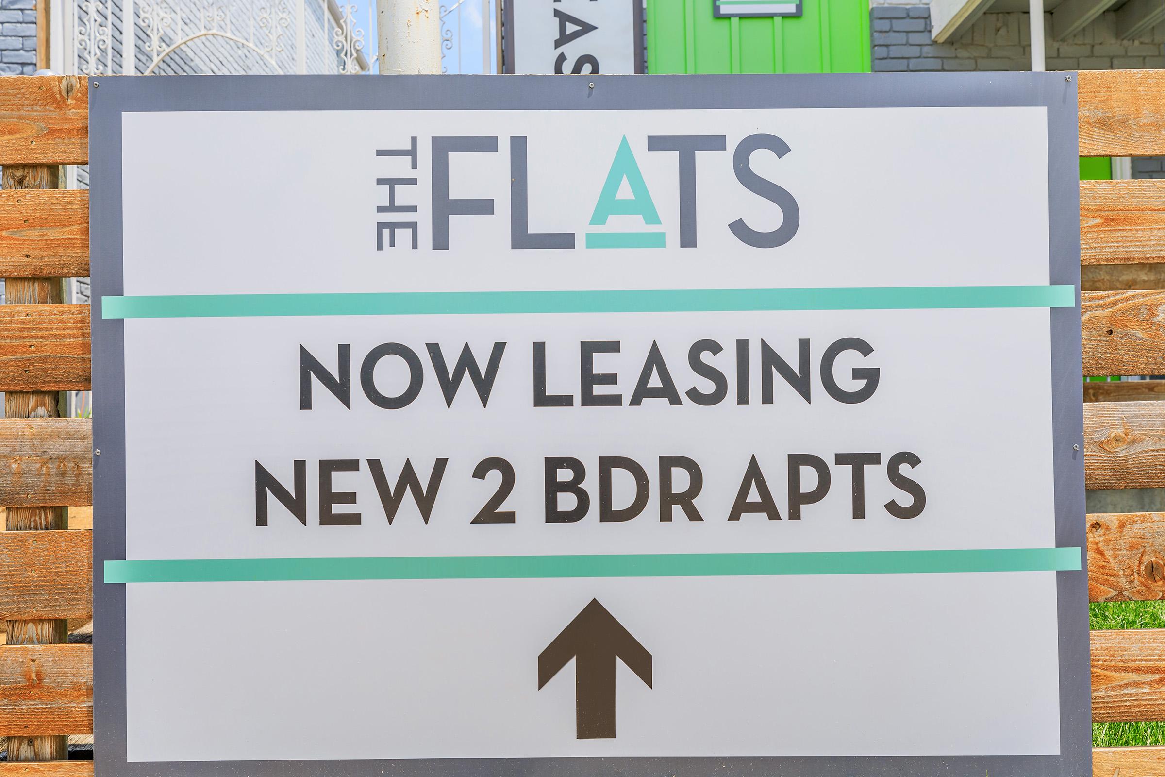 Sign advertising available apartments for rent, stating "The Flats" and "Now Leasing New 2 BDR Apts," with an upward-pointing arrow indicating direction.