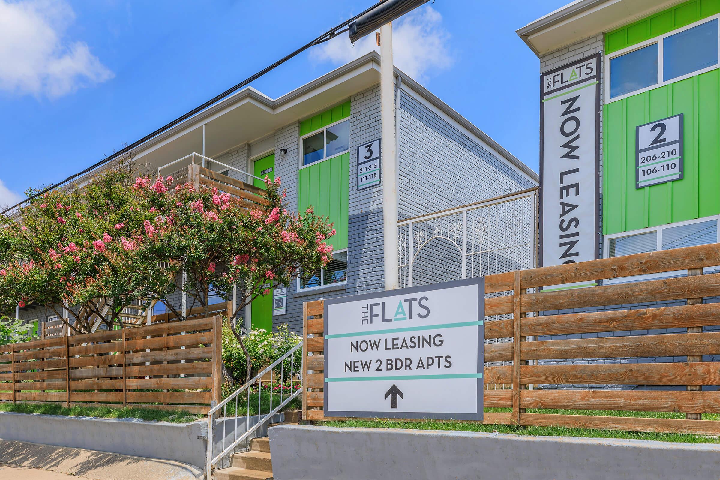 Modern apartment building with a green and white exterior. Features include multiple units with leasing signs, a wooden fence, flowering shrubs, and a clear sky. The sign in front indicates availability for new two-bedroom apartments.