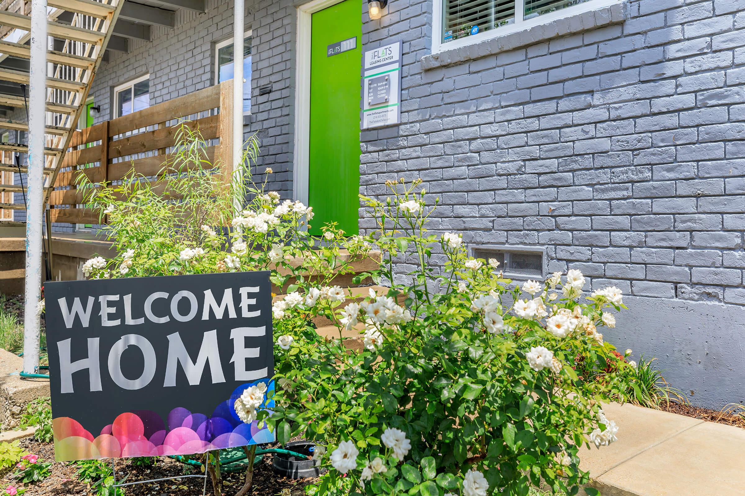 A bright green door is featured at the entrance of a building, surrounded by blooming white roses. In front, a large sign reads "WELCOME HOME" with colorful accents, set against a landscaped area. The building has a modern, welcoming appearance with a wooden fence and stairs leading up to the door.