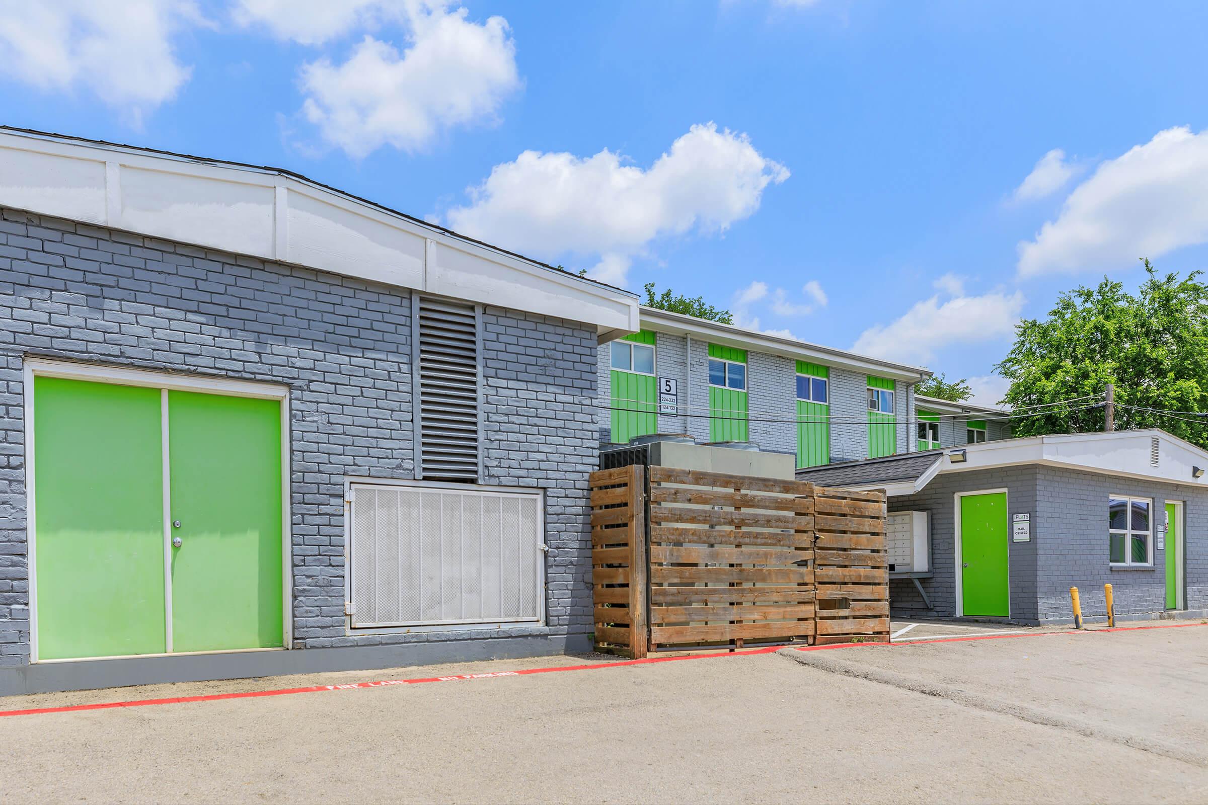 A modern building with gray brick walls and striking green doors and windows. In the foreground, a stack of wooden pallets is resting against the building. The scene is set under a bright blue sky with fluffy white clouds, showcasing a neat outdoor area.