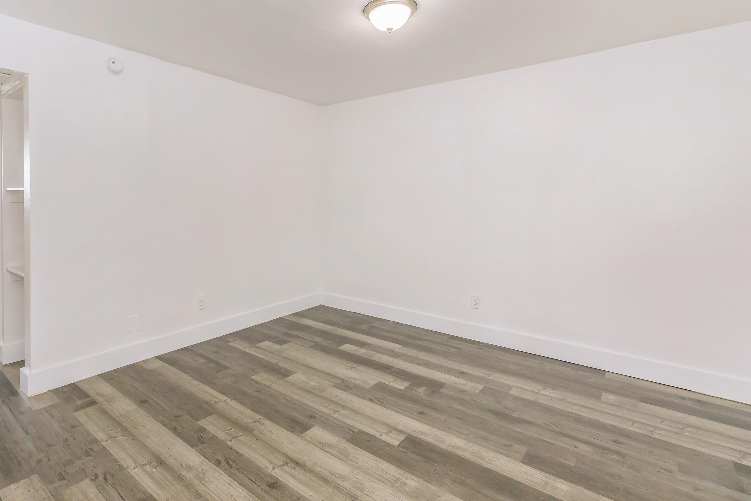 Empty room with white walls and a light-colored wood floor. A ceiling light fixture is visible, and there’s a small opening leading to another space on the left. The room appears fresh and clean, suitable for various uses or decoration.