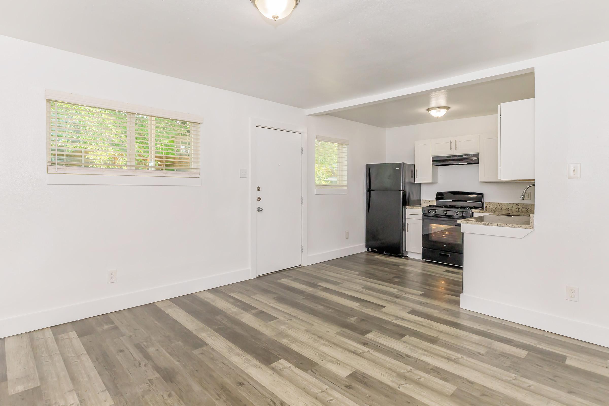 Bright and modern kitchen and living area with light wood-style flooring. Features white walls, a front door, a window with blinds, and an open kitchen with black appliances, including a refrigerator and a stove. Natural light filters in, creating a welcoming atmosphere.