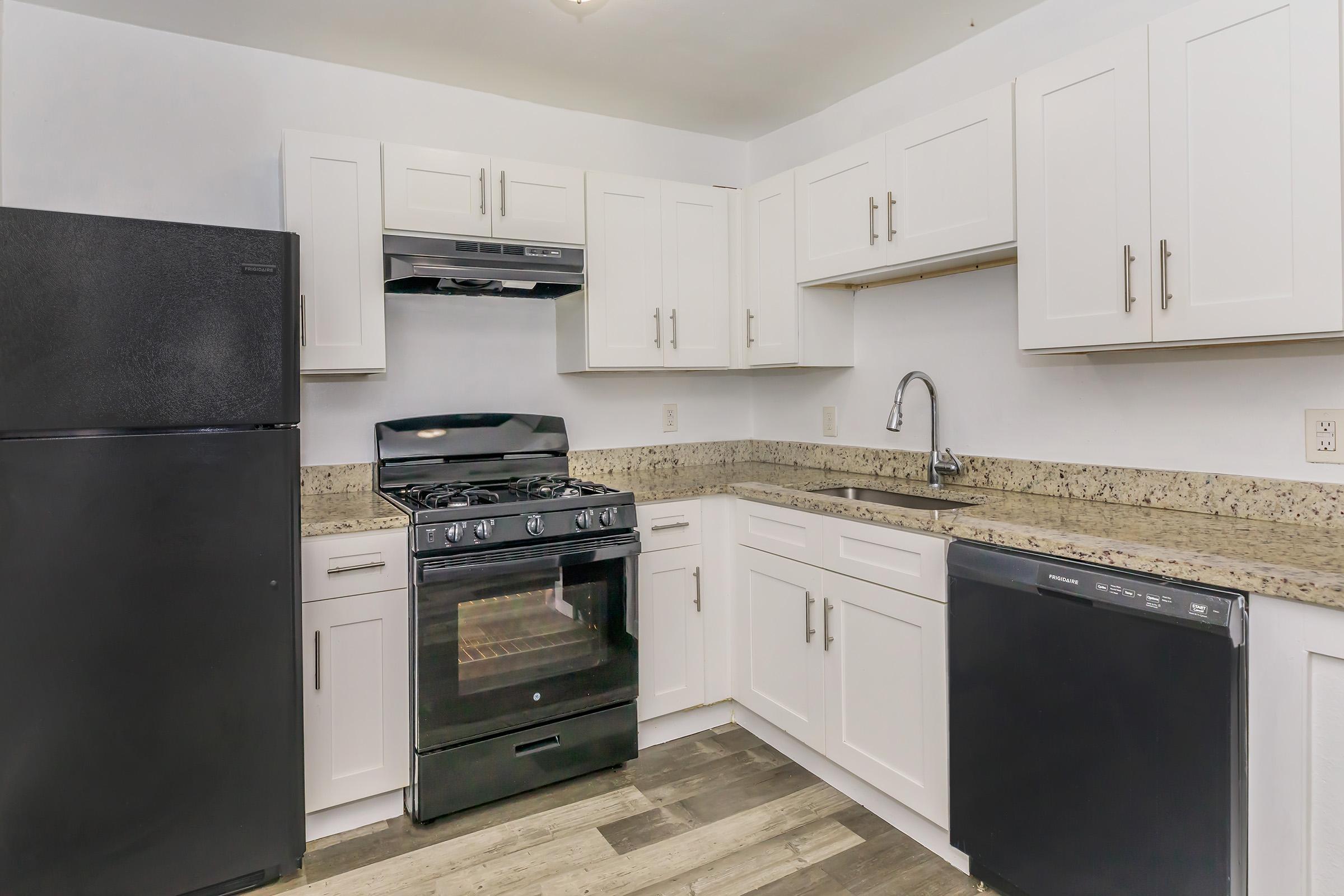 Modern kitchen featuring white cabinetry, stainless steel appliances including a gas stove, refrigerator, and dishwasher. The countertops are made of speckled granite, and the floor is a mix of wood-like tiles. Natural light is provided by overhead lighting.