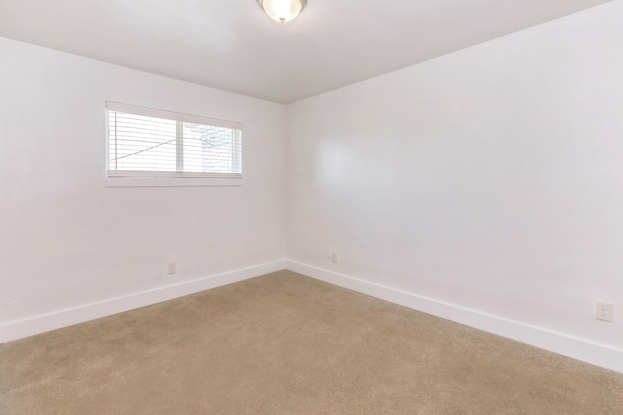 Empty room with light-colored walls and beige carpeting. A small window with blinds allows natural light to enter. The ceiling is plain with a light fixture. The space is uncluttered and minimalist, suggesting a fresh start or potential for customization.