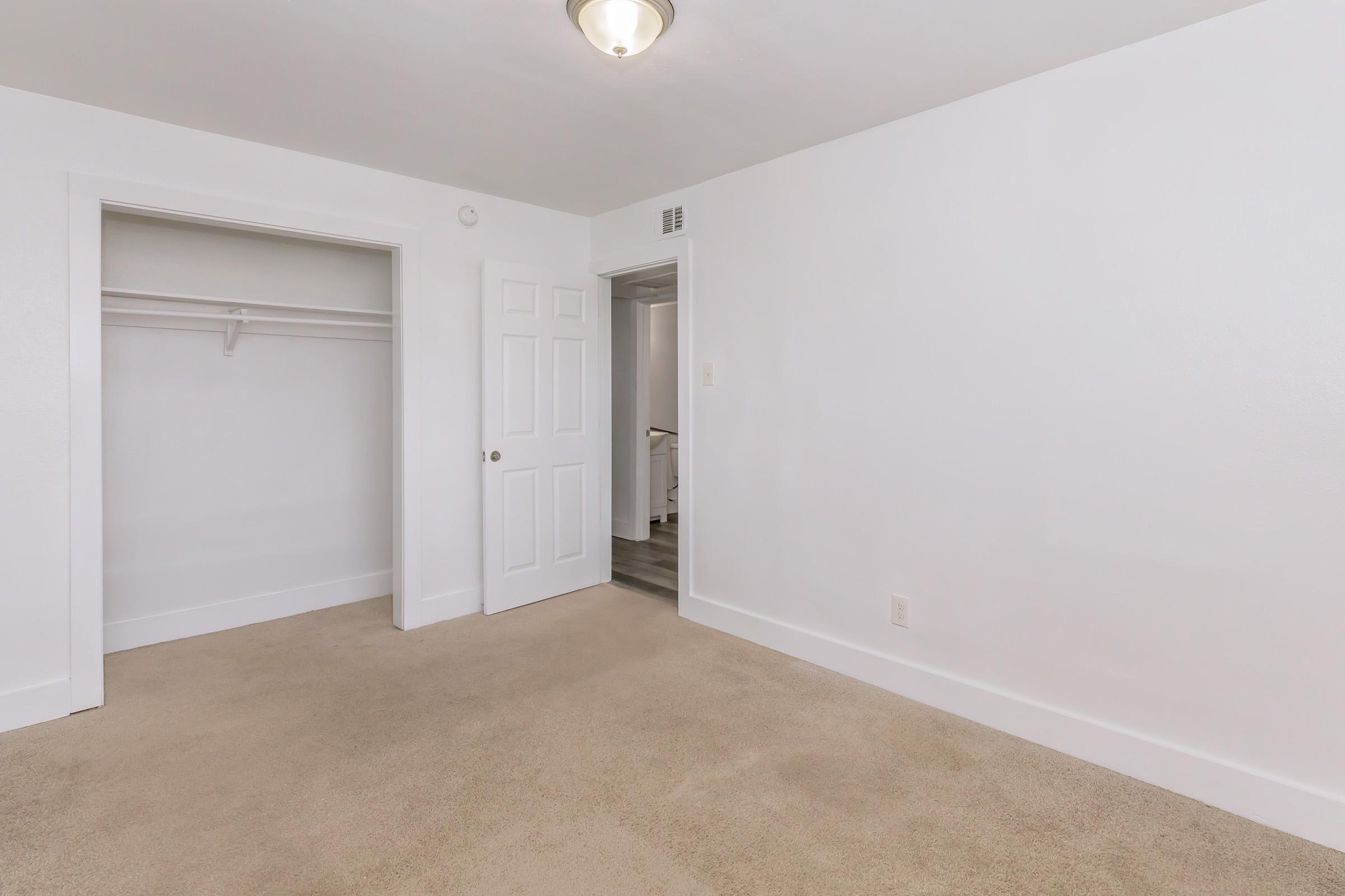 A bright, empty room with white walls and a light fixture on the ceiling. The floor is carpeted in a neutral color. One wall features a doorway leading to another room, while another wall has a closet with sliding doors. The overall atmosphere is clean and minimalistic, suggesting potential for various uses.