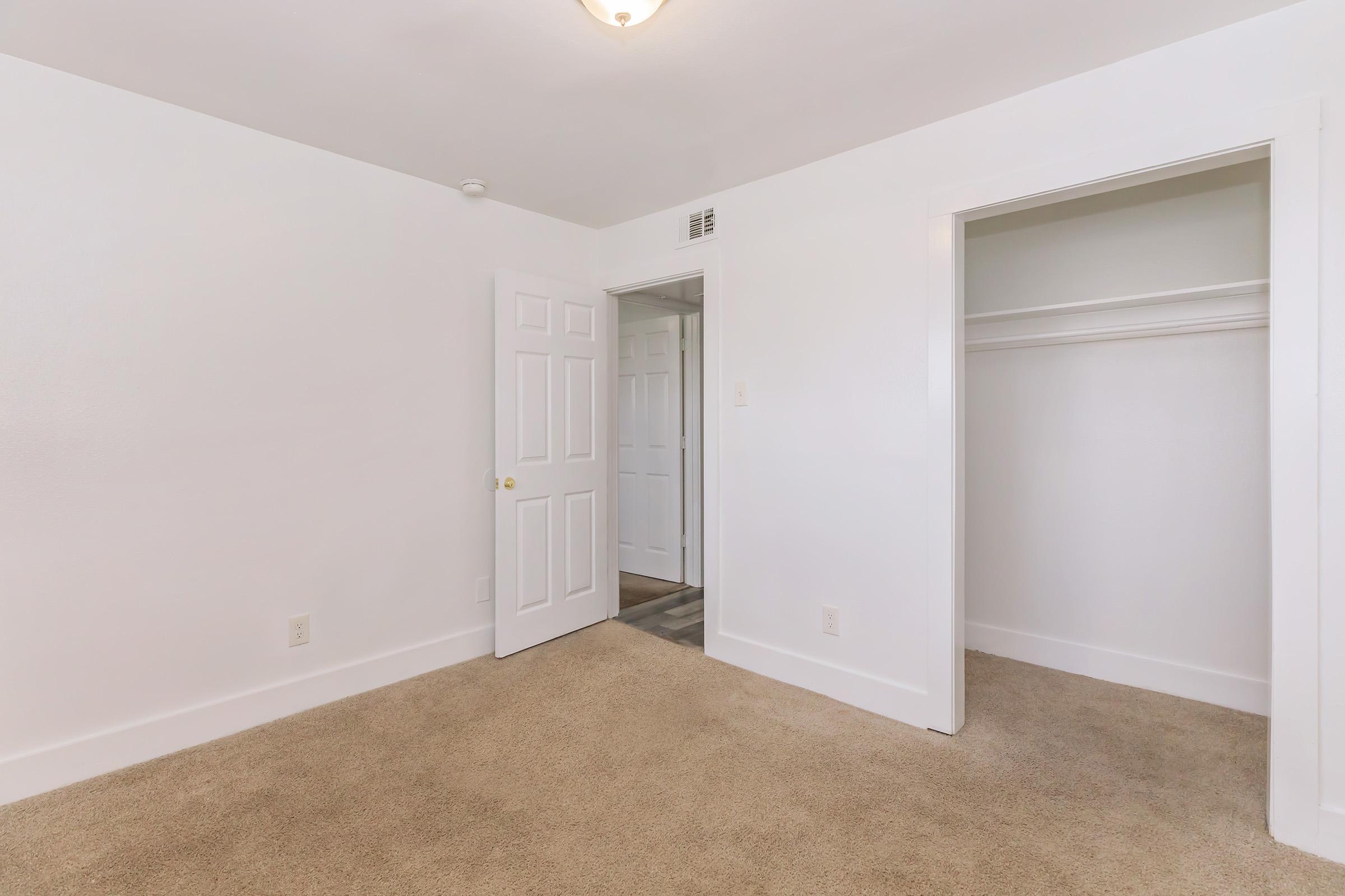 Empty room with light beige carpet flooring, white walls, and a ceiling light fixture. A closed door leads to another space, and there is a closet with white shelving in the corner. The room has a clean and modern look, making it suitable for various purposes.
