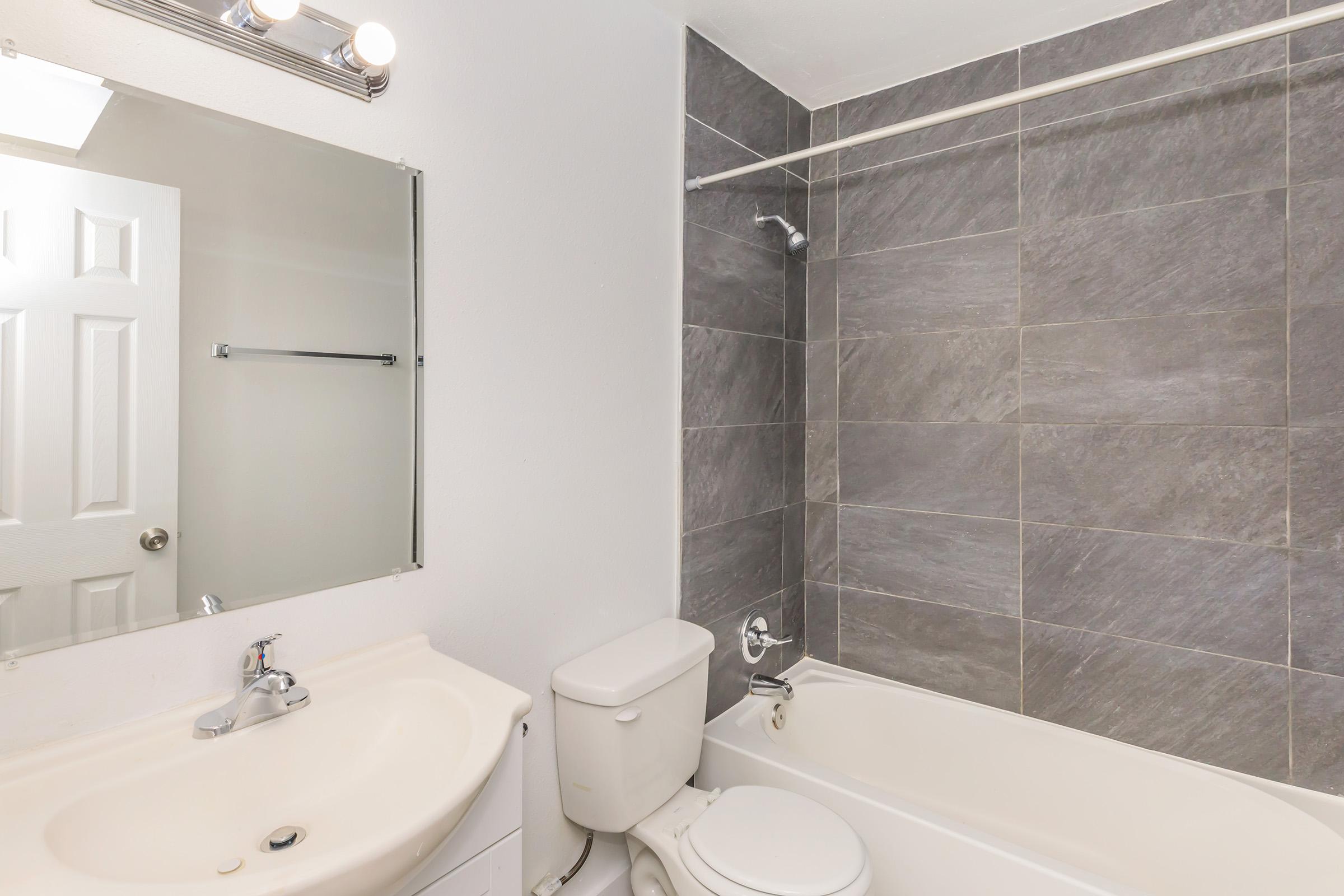 A clean bathroom featuring a white bathtub with a showerhead, a white toilet, and a sink with a modern faucet. The walls are covered in dark gray tiles, and there is a mirror above the sink. A door is visible in the background, leading to another area.