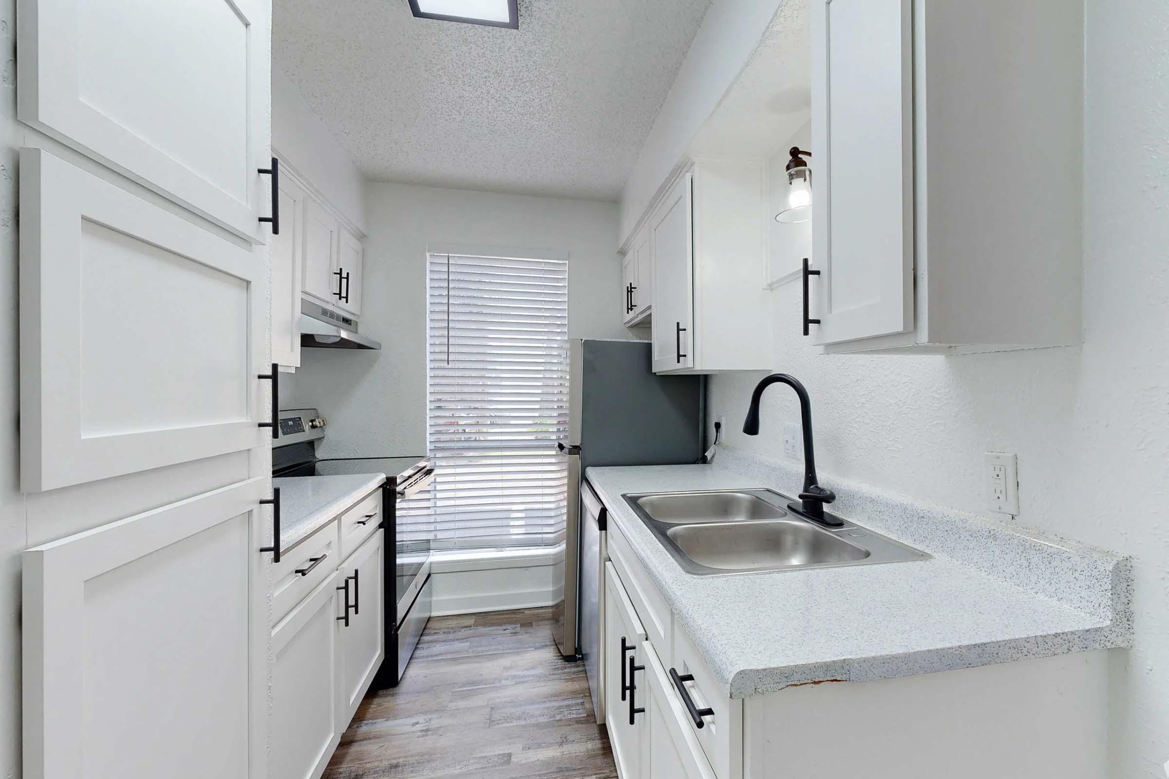 Modern kitchen featuring white cabinetry, a dual sink with a black faucet, and stainless steel appliances. Natural light enters through a window with blinds, illuminating the clean design and granite countertops. The flooring is a light wood finish, contributing to a bright and inviting atmosphere.