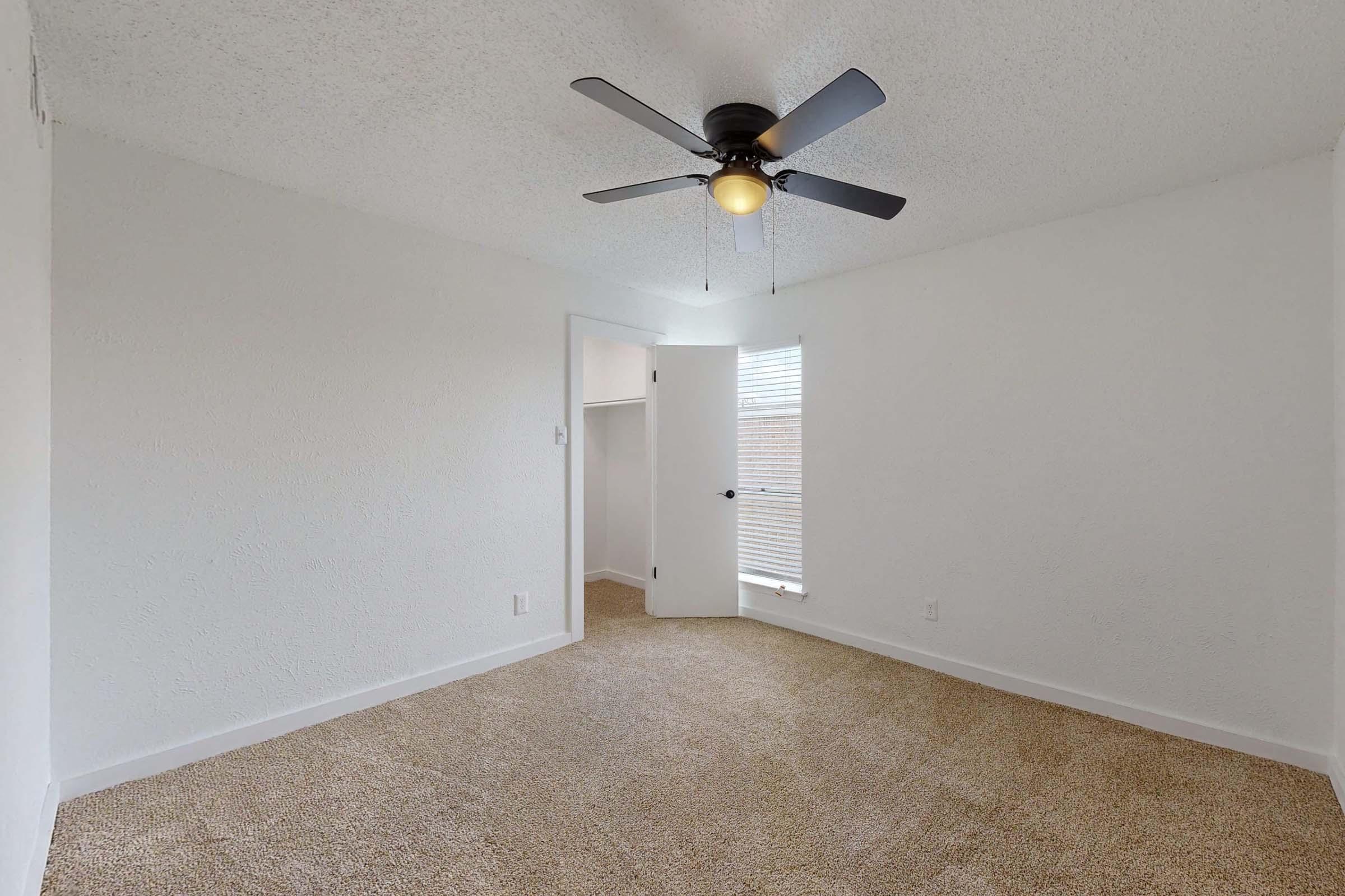 A bright, empty room with a light brown carpet and white walls. It features a ceiling fan with three blades and light, a door leading to another space, and a window with blinds allowing natural light to enter. The room has a spacious feel with no furniture present.