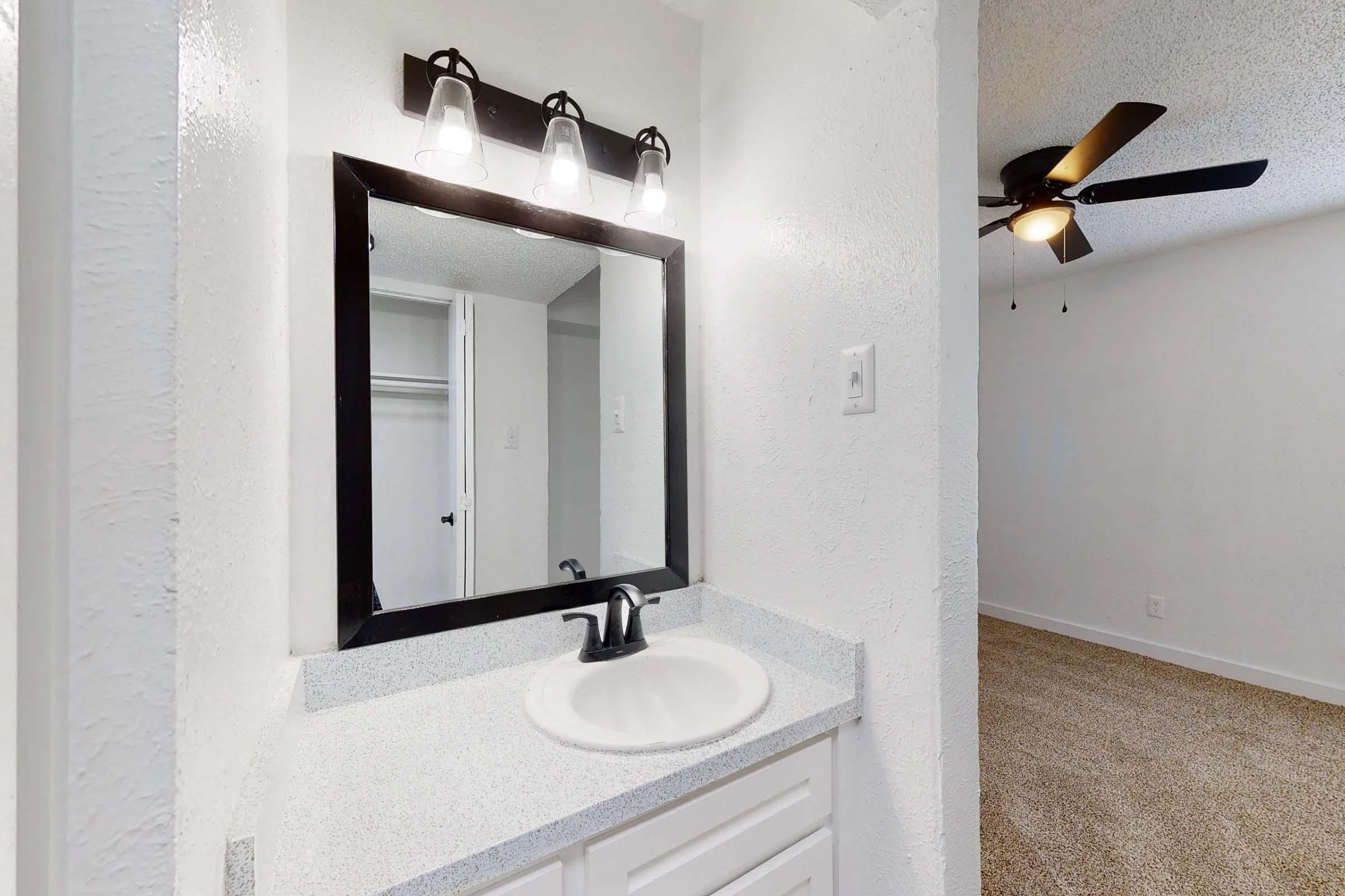 A modern bathroom featuring a white sink and a rectangular mirror above it, illuminated by three stylish light fixtures. The wall is painted white, with a glimpse of a closet space on the left. A ceiling fan is visible in the background against a light-colored ceiling. The space appears bright and well-maintained.