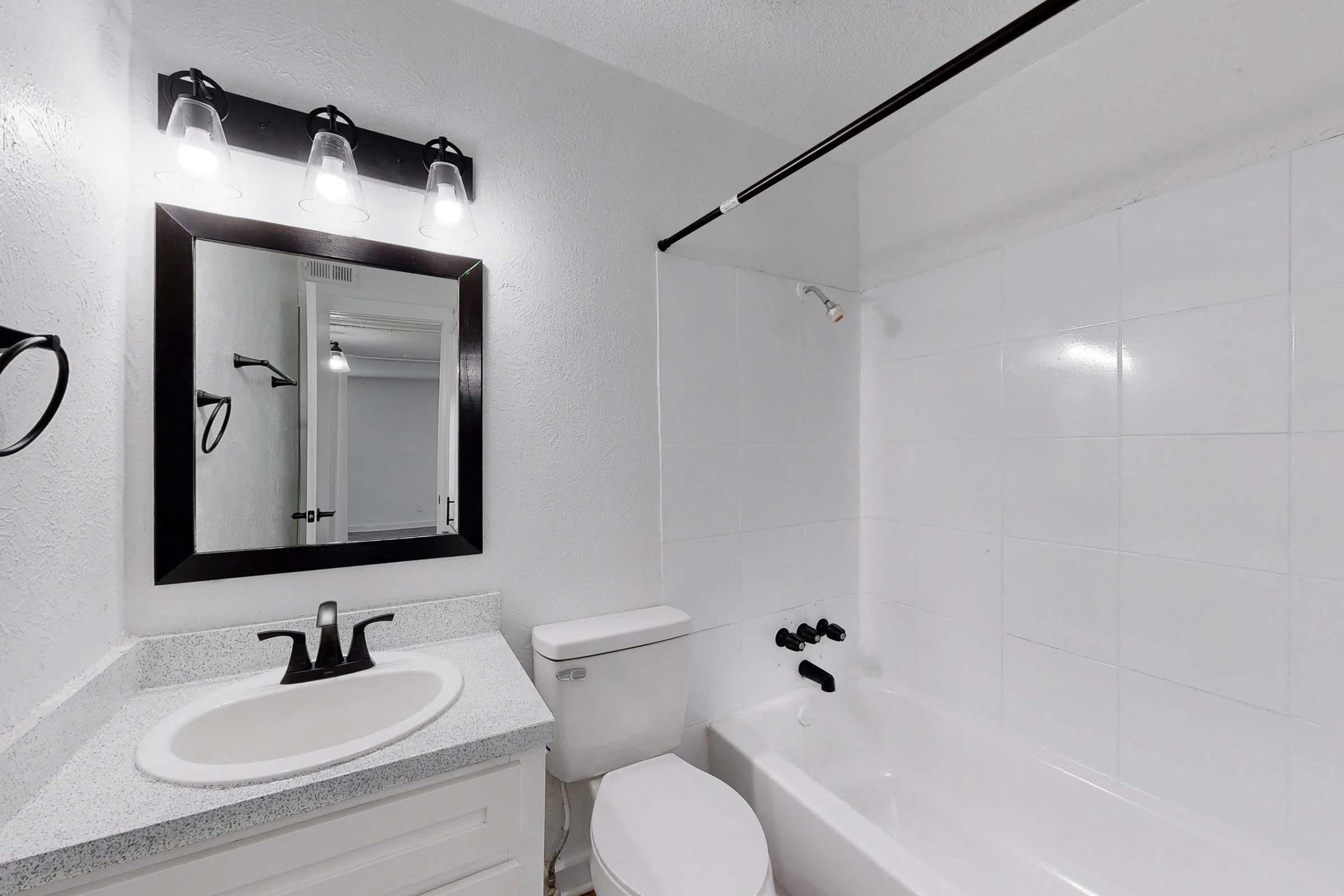 A clean, modern bathroom featuring a white bathtub, beige countertop with a sink, a black faucet, and a mirror framed in black. The walls are tiled in white, and there is a shower rod above the tub with a showerhead. Bright lighting is provided by four fixtures above the mirror.