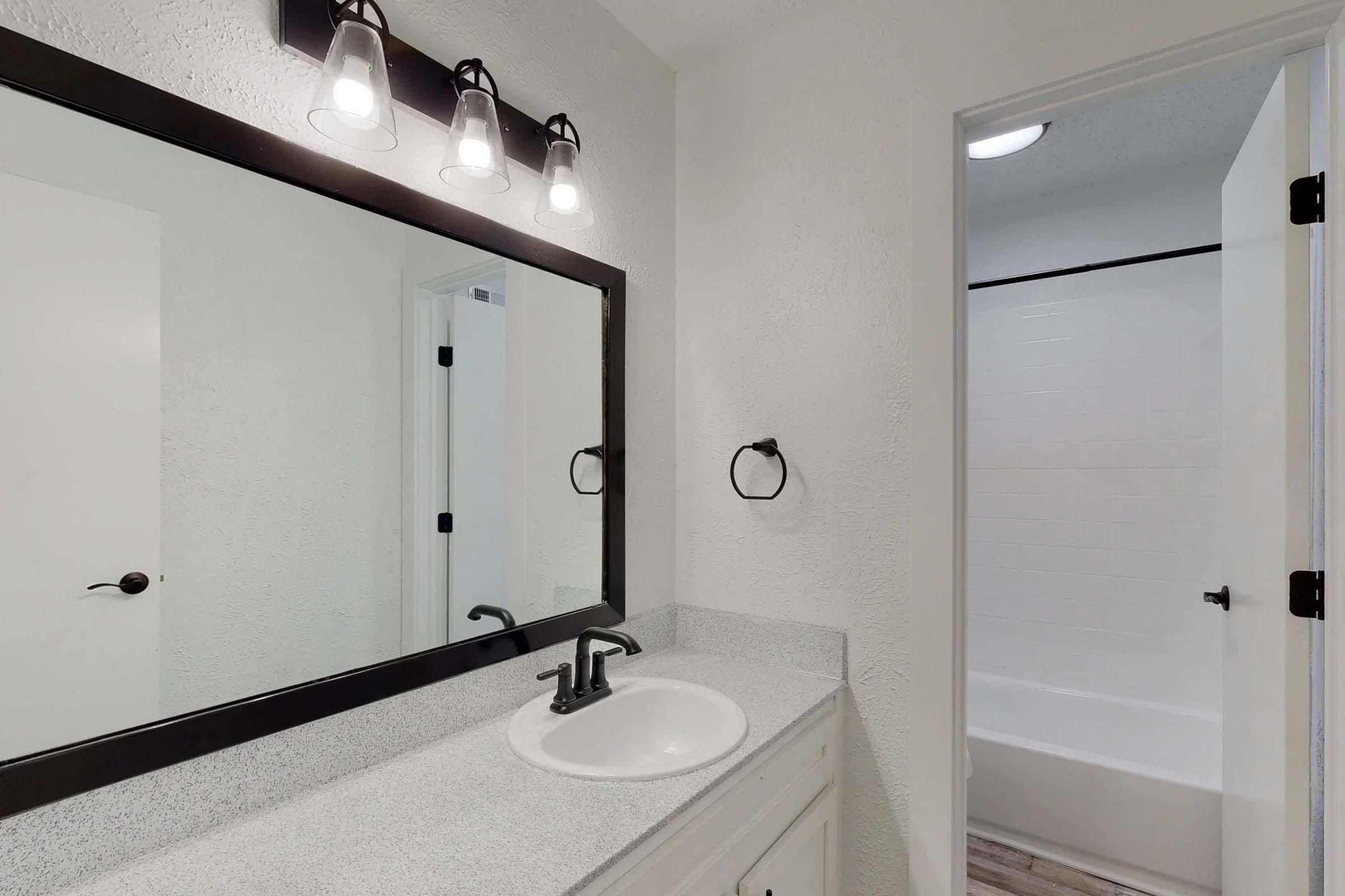 A modern bathroom featuring a white painted wall, a large mirror above a countertop with a sink and faucet, three pendant lights above the mirror, and a towel holder. In the background, a door leads to a separate area with a bathtub surrounded by white tile.