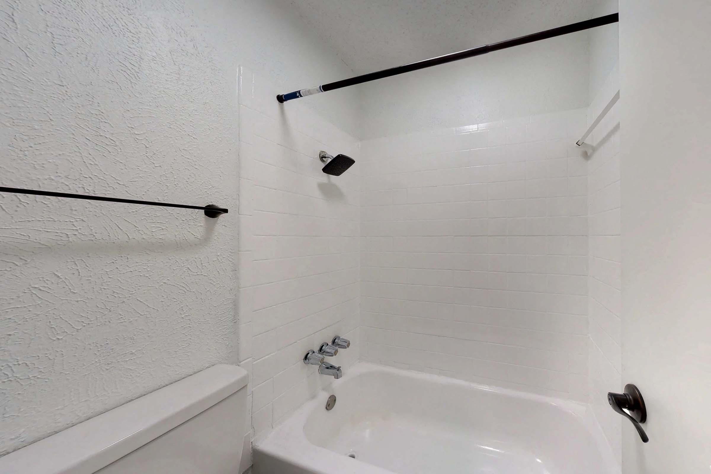 A simple bathroom with a white tiled shower area, featuring a showerhead, a bathtub, and a toilet. The walls are textured white, and there are two black towel bars mounted on the walls. The space is clean and well-lit, showcasing a minimalistic design.