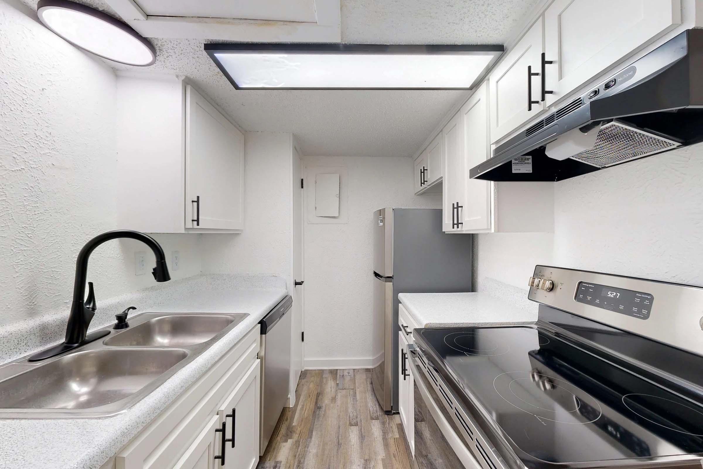 Modern kitchen featuring white cabinetry, stainless steel appliances, a double sink, and an electric stove. The space has a sleek design with a light countertop and wood-look flooring, illuminated by overhead lighting and a ceiling fixture.