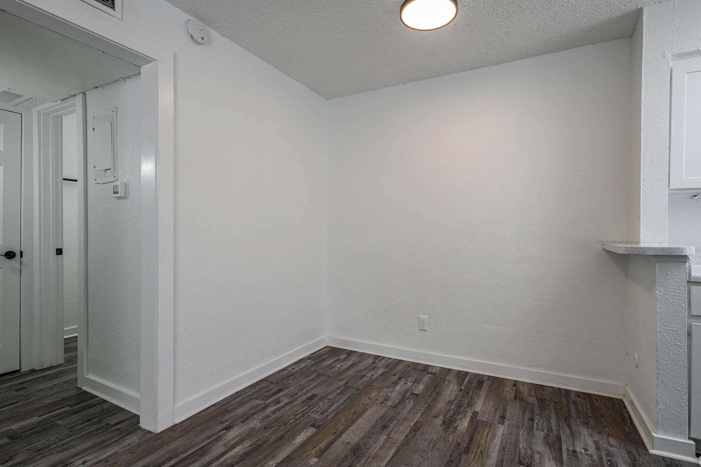 A lightly colored, empty room with a textured white wall and a ceiling light fixture. The floor is covered with brown, wood-like laminate. To the left, there is an open doorway leading to another room, and a small counter visible on the right side, indicating a kitchen or dining area.