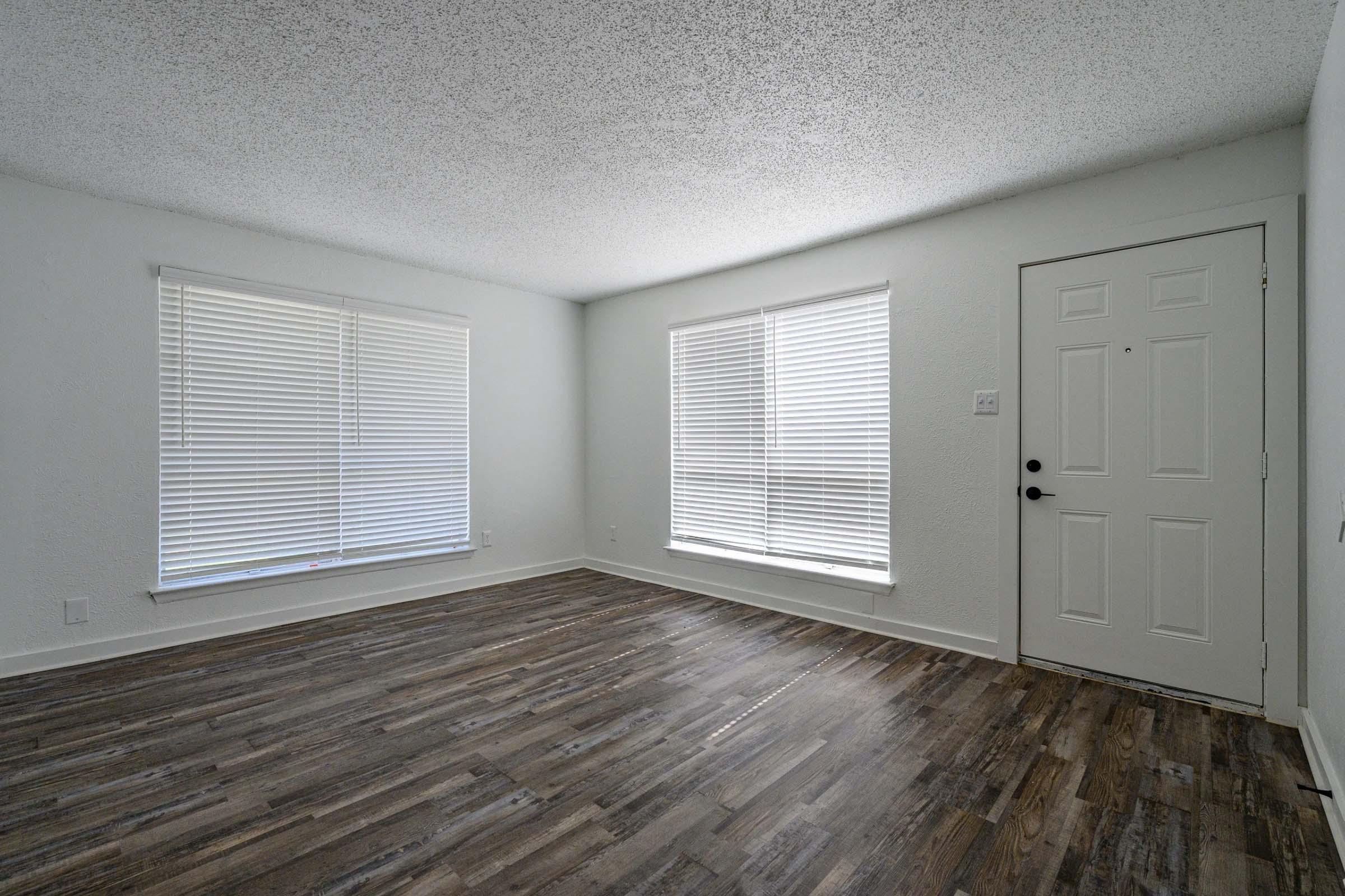 Empty room with light-colored walls and wooden flooring. There are two windows with white blinds allowing natural light, and a front door is visible on the right side. The space is clean and uncluttered, ideal for various interior design possibilities.