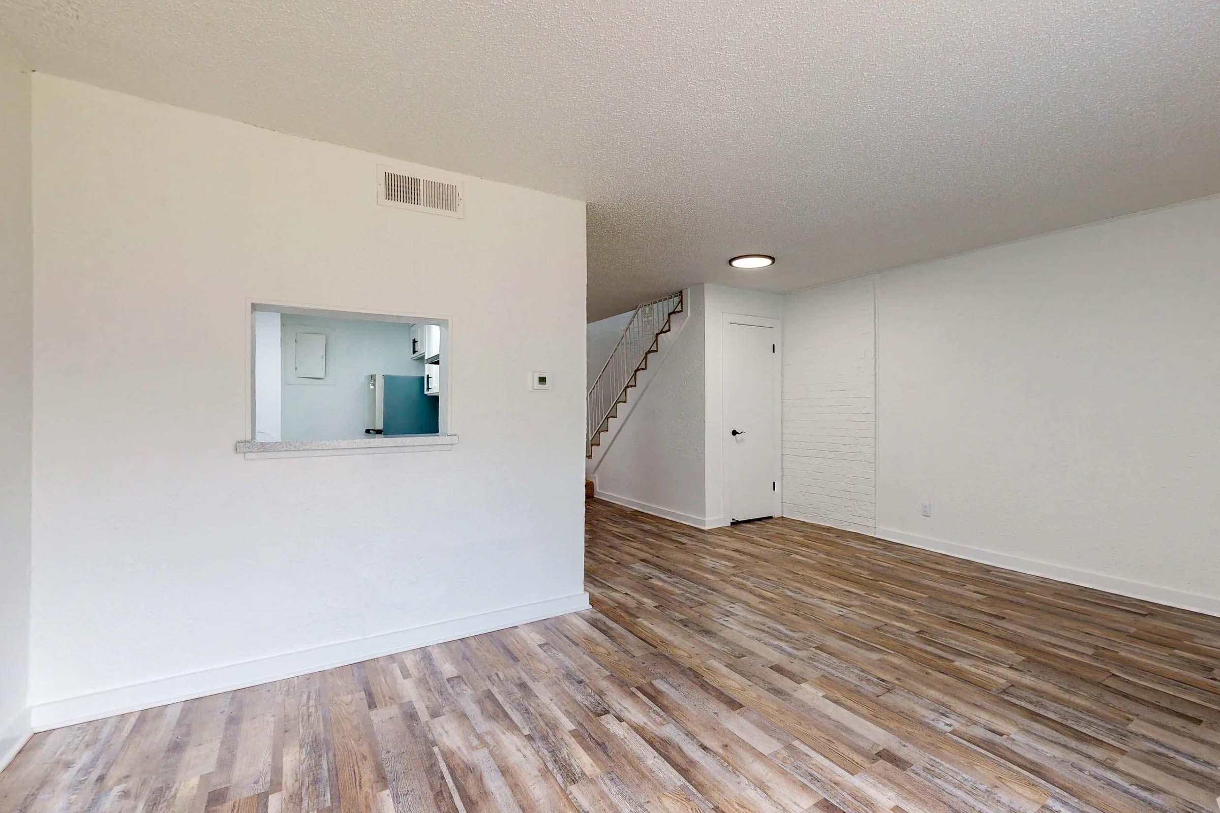 Interior view of a bright, airy room with white walls and wooden flooring. A small pass-through window leads to a kitchenette area visible in the background. To the right, there is a stairway leading to an upper level, and the space has a modern, minimalist design.