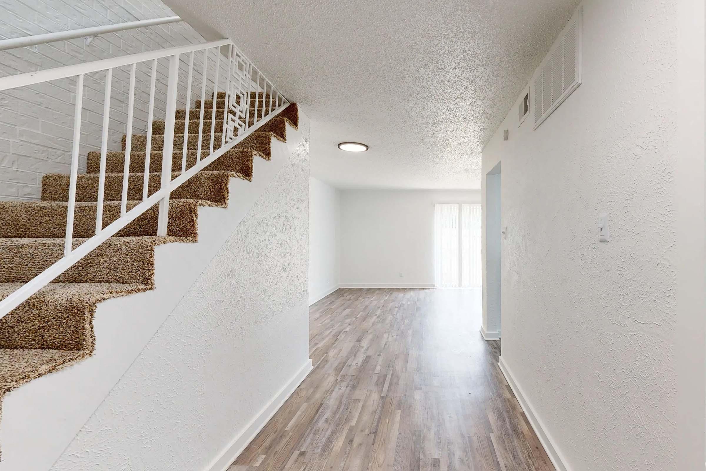 Interior view of a spacious hallway featuring a carpeted staircase on the left, light-colored walls, and laminate flooring. A round ceiling light illuminates the area, while natural light enters from a sliding door at the far end. The space appears clean and modern, with a minimalist design.