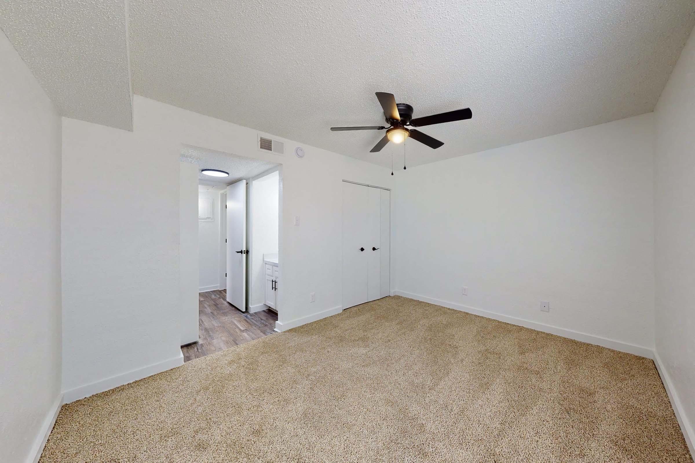A spacious, well-lit bedroom featuring a ceiling fan and a soft carpeted floor. The walls are painted white, and there is a door leading to a bathroom area in the background. Natural light enters from a window, creating a cozy atmosphere.