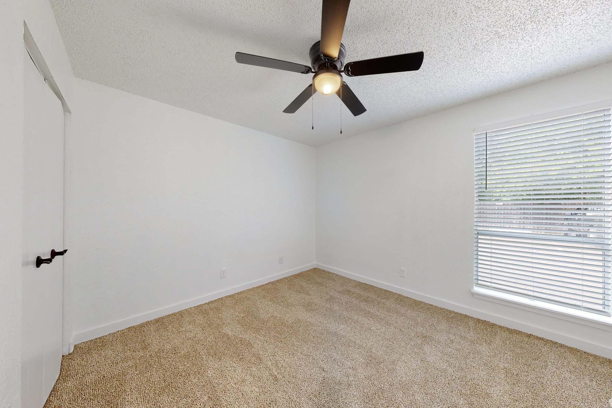 A bright, empty room with light-colored walls and a textured ceiling. It features a ceiling fan with a light fixture, a window with blinds allowing natural light in, and carpeted flooring. The space has a minimalist design, ideal for various furnishings and personal touches.