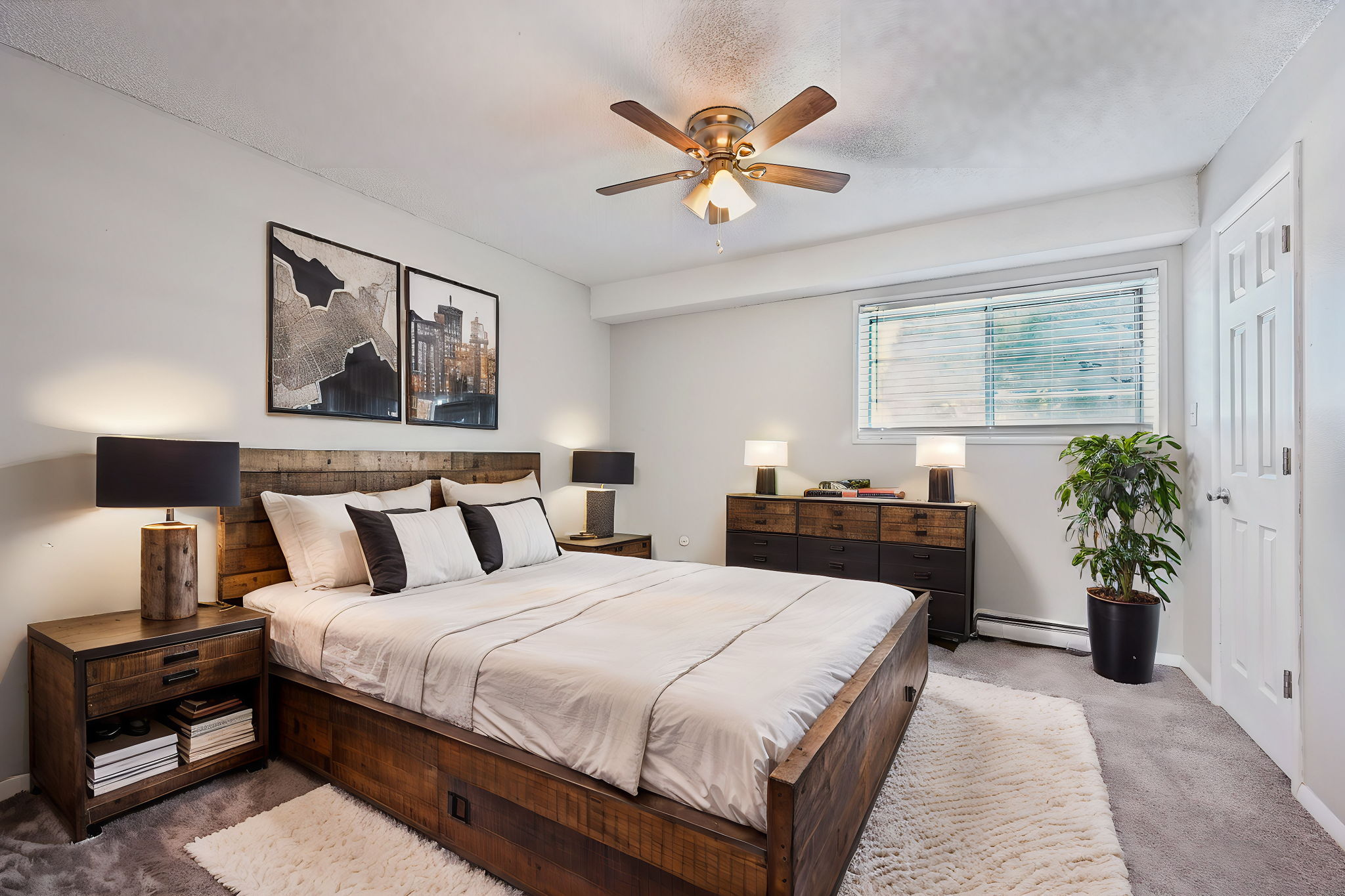 A modern bedroom featuring a large bed with white bedding and decorative pillows. There are two framed artworks on the wall, a ceiling fan, and bedside lamps on wooden nightstands. A wooden dresser is positioned against the wall, and a small plant is situated in the corner near a window with natural light.