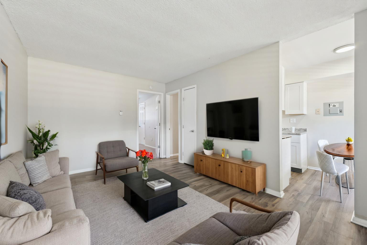 A modern living room featuring a light-colored sofa and an armchair, with a coffee table in the center. A large flat-screen TV is mounted on the wall, and a wooden media unit holds decorative items. A dining area with a round table is visible in the background, along with a kitchen area. Flowers add a touch of color.