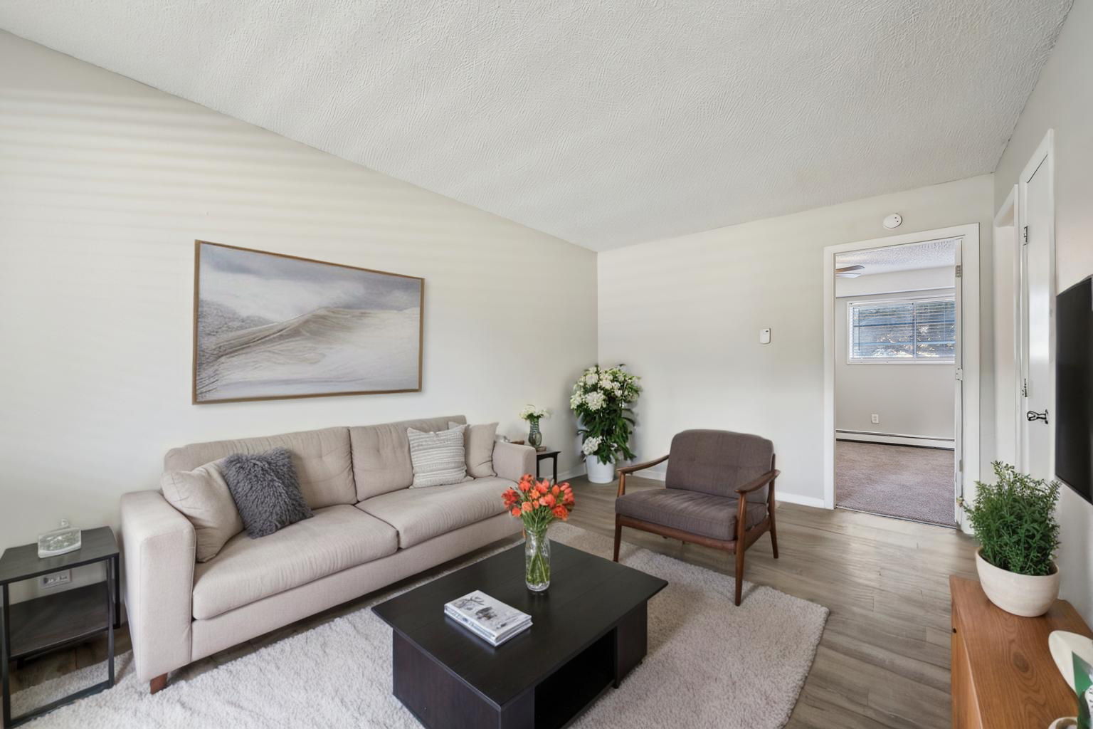 A cozy living room featuring a light gray sofa with decorative pillows, a dark coffee table with a vase of orange flowers, and a comfortable chair. A large painting hangs on the wall, and a potted plant adds greenery. A doorway leads to another room, with natural light filtering through a window.