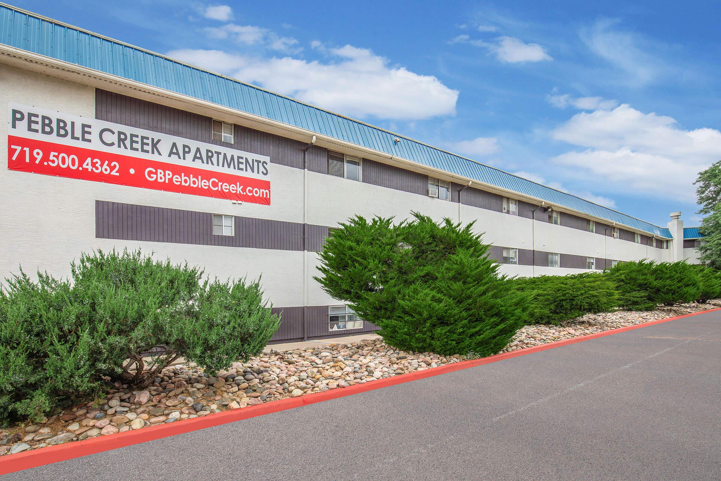 A view of the Pebble Creek Apartments exterior, showcasing a two-story building with a blue roof. Various shrubs and rocks line the pathway, and a large sign with the apartments' name, phone number, and website is prominently displayed on the building's side. Clear blue skies can be seen in the background.