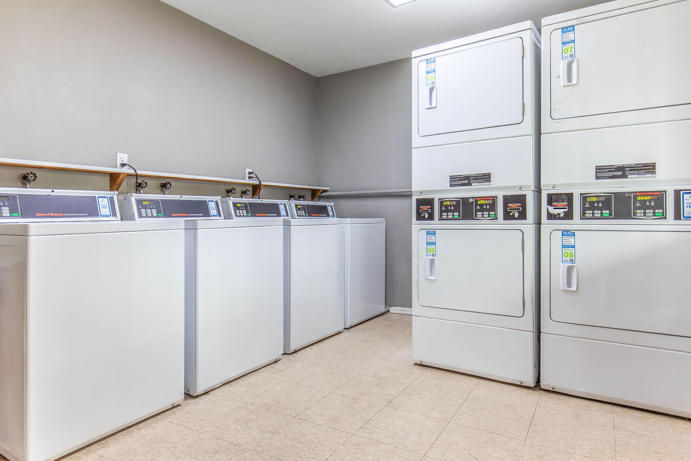 A clean, well-lit laundry room featuring multiple stacked washing machines and dryers, with a neutral wall color and tiled floor. The appliances have digital controls, and the space is organized with shelves above.