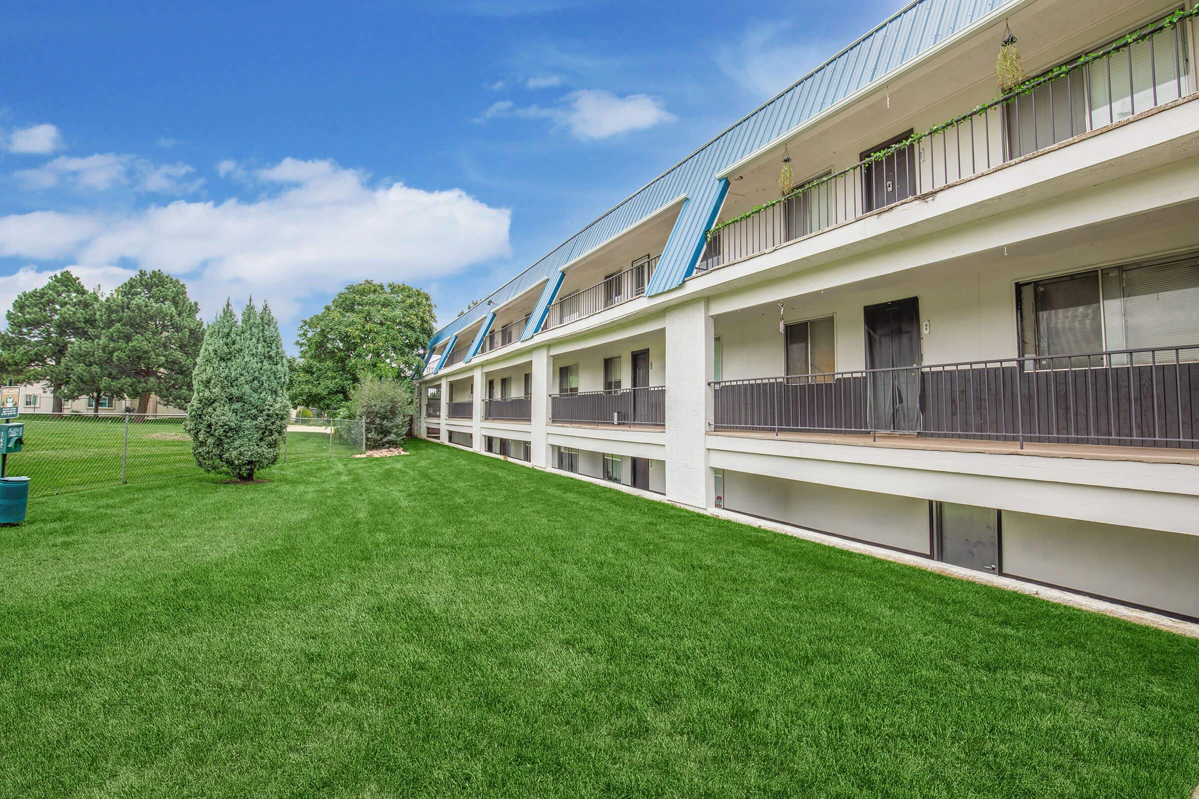 A well-maintained, two-story building with a white exterior and blue accents. The structure features multiple balconies and overlooks a lush green lawn. Amidst the well-manicured grass, there are trees, creating a tranquil outdoor environment. The sky is bright with a few clouds, adding to the serene atmosphere.