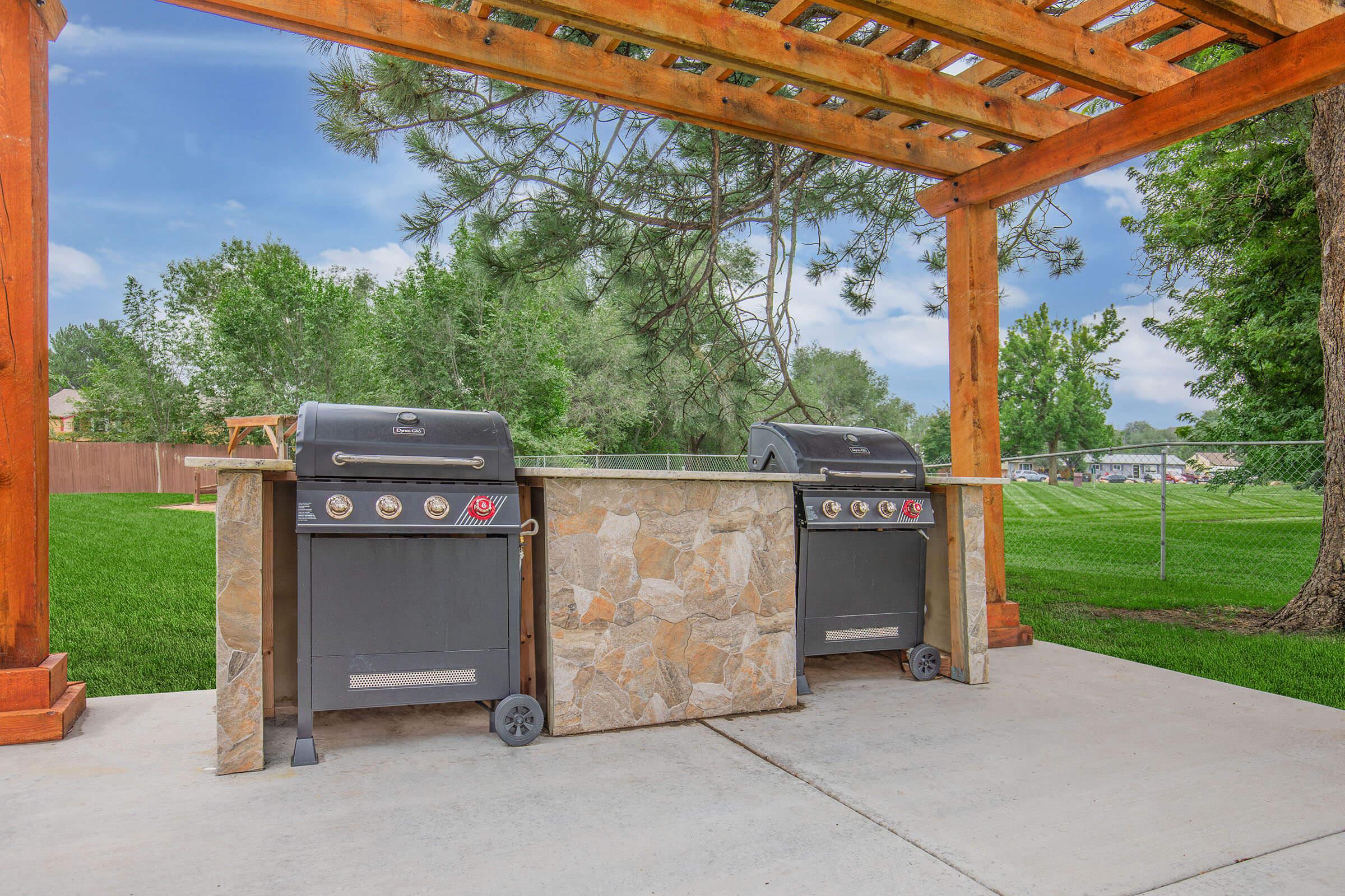 Outdoor grilling station featuring two black gas grills set on a stone island, surrounded by a green lawn and trees. A wooden pergola overhead provides shade, and a chain-link fence encloses the area in the background. The scene is bright and inviting, ideal for outdoor cooking and gatherings.