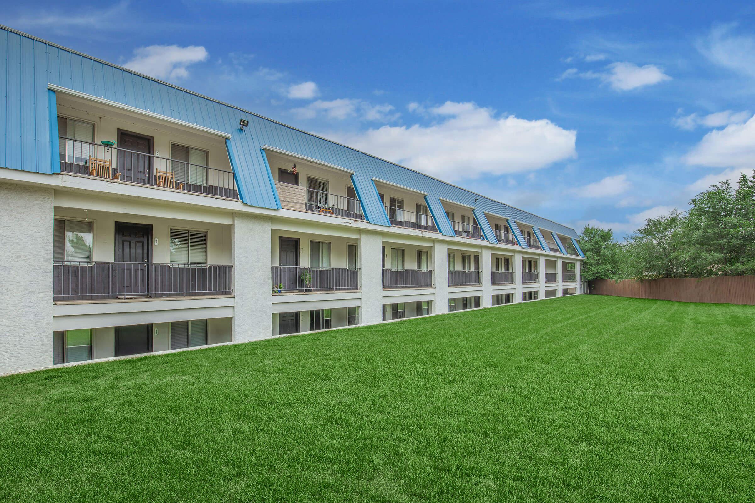 A modern multi-story building with a light-colored exterior and a blue roof, featuring several balconies. The building is set against a bright sky with some clouds, and a neatly manicured green lawn in the foreground, providing a welcoming appearance.