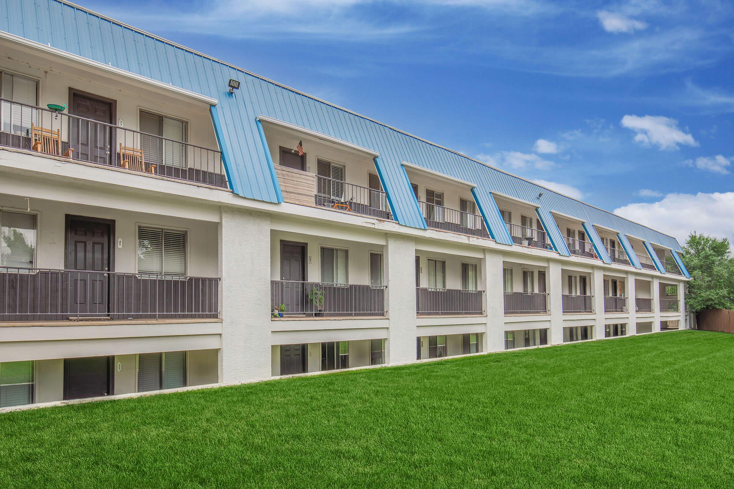 A modern two-story building featuring multiple balconies with blue roofs, surrounded by green grass and a clear blue sky. The building has a clean, simple design, with large windows and sliding doors. The landscaping is well-maintained, creating an inviting atmosphere.
