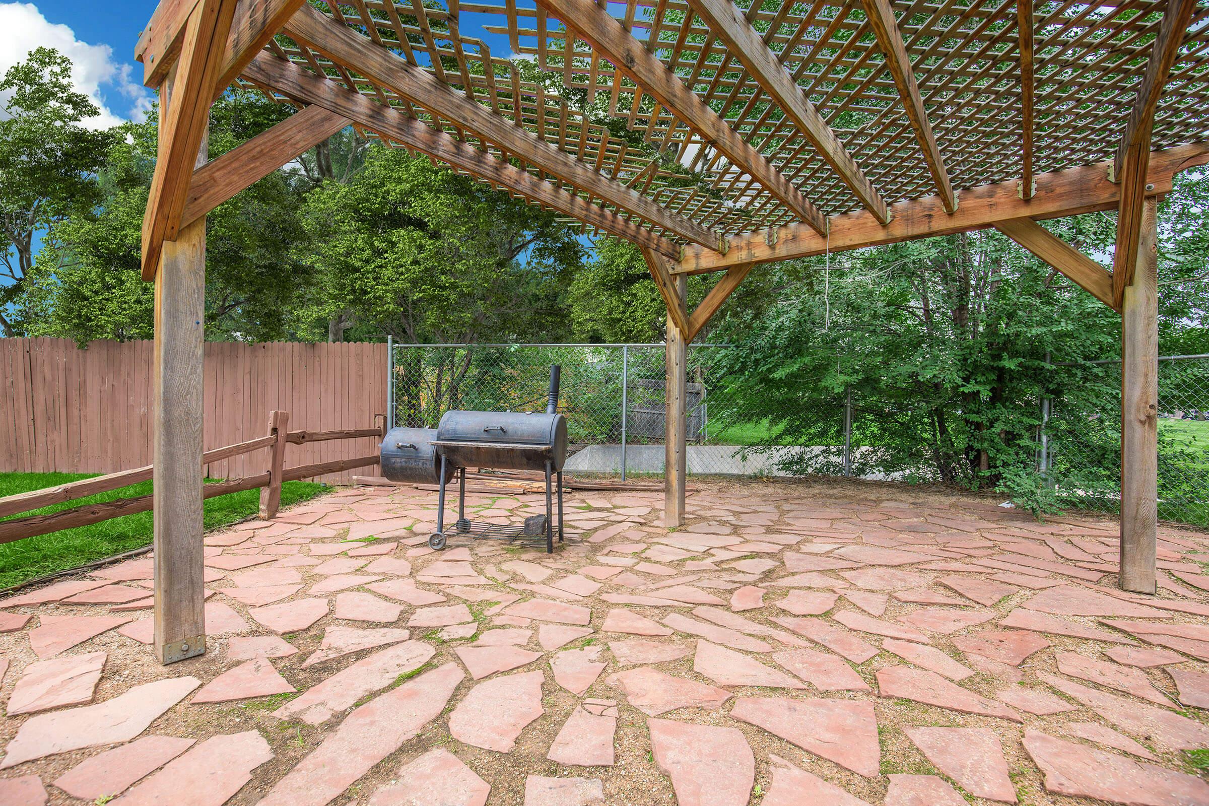 A rustic outdoor patio with a stone floor and wooden pergola, featuring a barbecue grill at the center. Lush greenery surrounds the area, and a wooden fence is visible in the background, creating a peaceful and inviting space for outdoor cooking and gatherings.