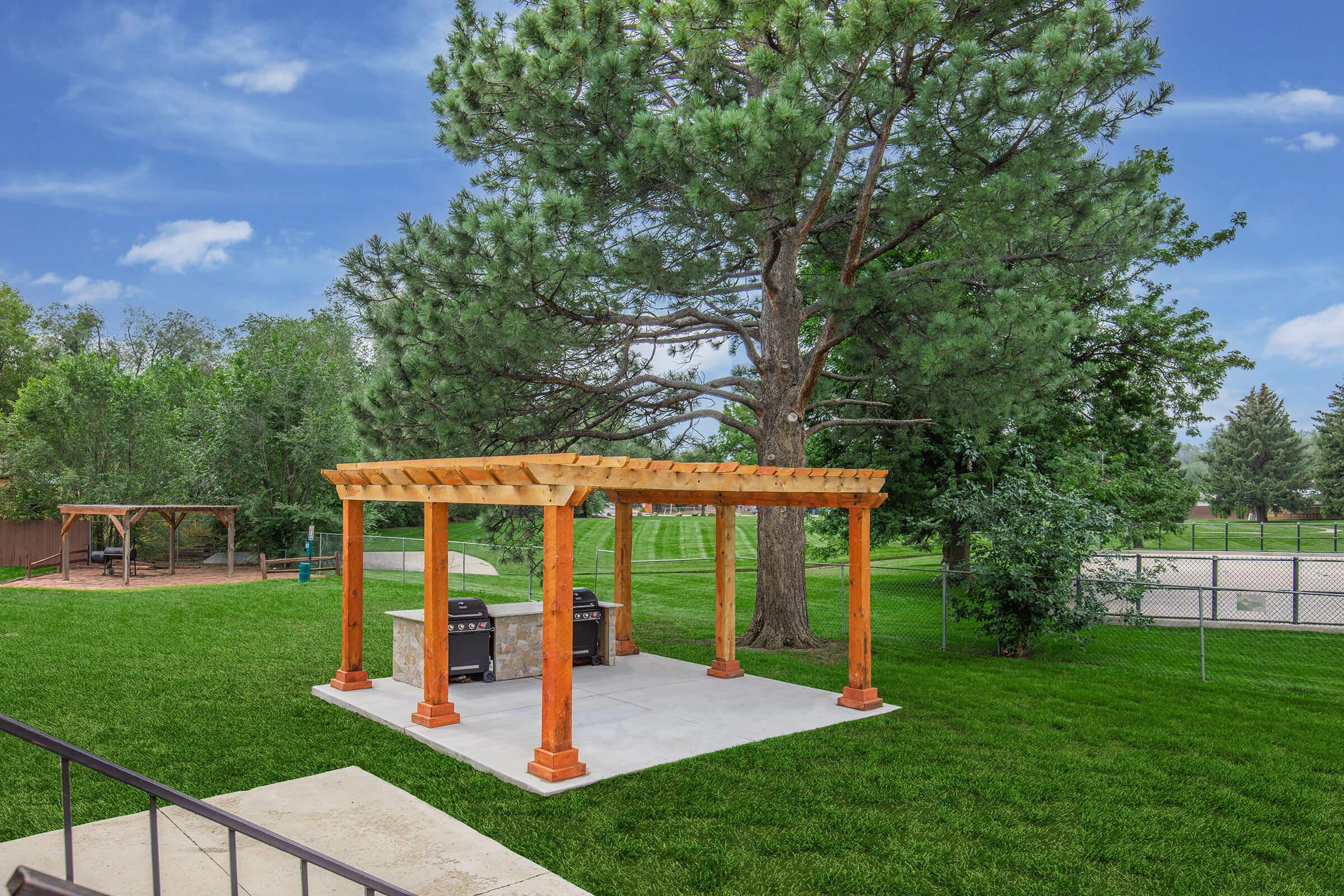 A spacious backyard scene featuring a wooden pergola with a concrete base, set in a lush green lawn. Nearby, a large tree stands tall, and in the background, there are recreational facilities, including a playground and a fenced area. The sky is clear with a few clouds, enhancing the inviting atmosphere.