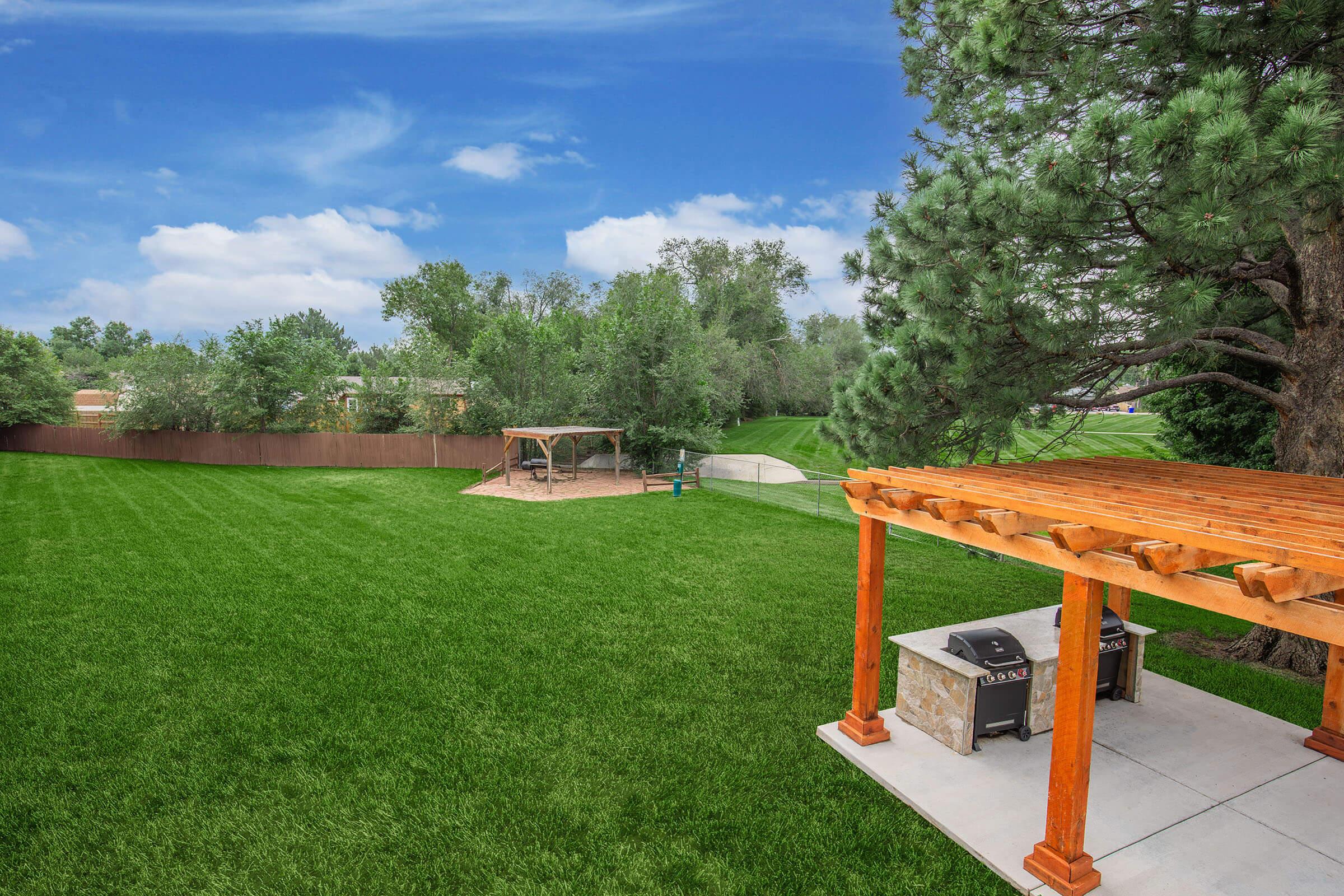 A spacious backyard featuring a lush green lawn, a wooden pergola, and a concrete patio. In the background, there is a play area with swings and a slide, surrounded by trees and a fence, under a blue sky with scattered clouds. Perfect for outdoor activities and relaxation.