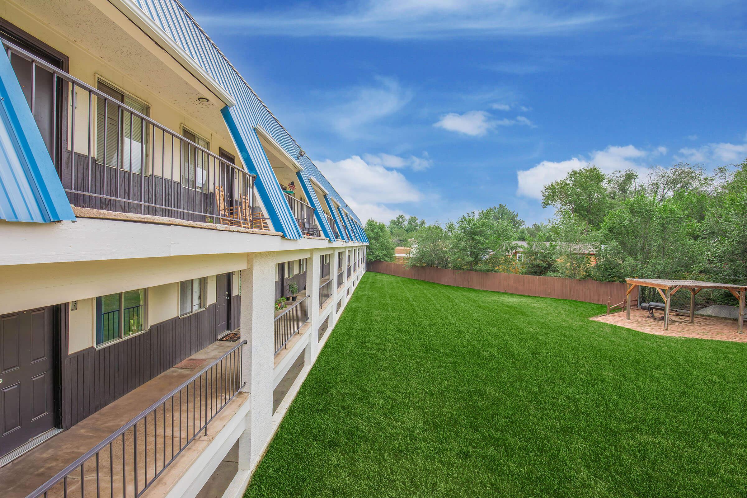 Exterior view of a two-story motel with blue accents and balconies. A well-maintained grassy area is visible in the foreground, surrounded by trees and a wooden fence. The sky is clear with a few clouds, creating a bright and inviting atmosphere.