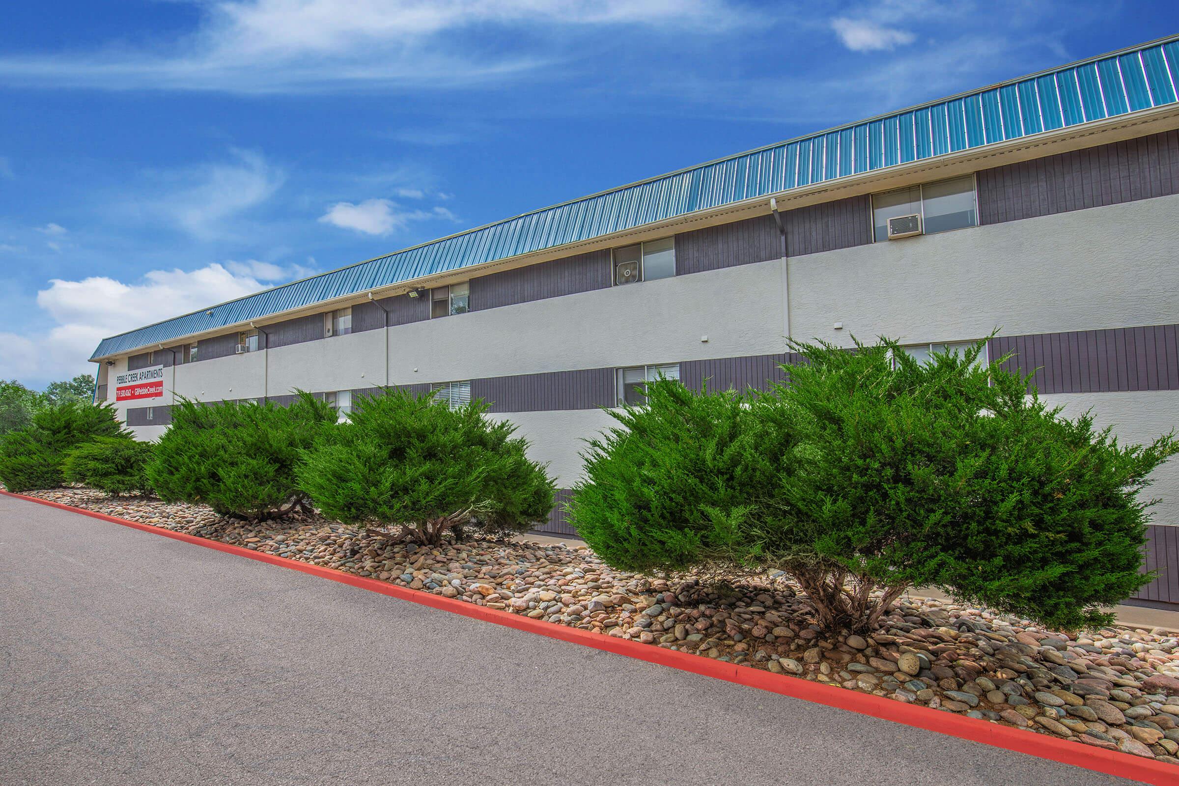 A modern two-story building with a blue metal roof, featuring large windows and a smooth exterior. Neatly trimmed shrubs line the pathway leading to the entrance, which is marked by a sign. The sky is clear with a few clouds, and the area is well-maintained with decorative stone landscaping along the edges.