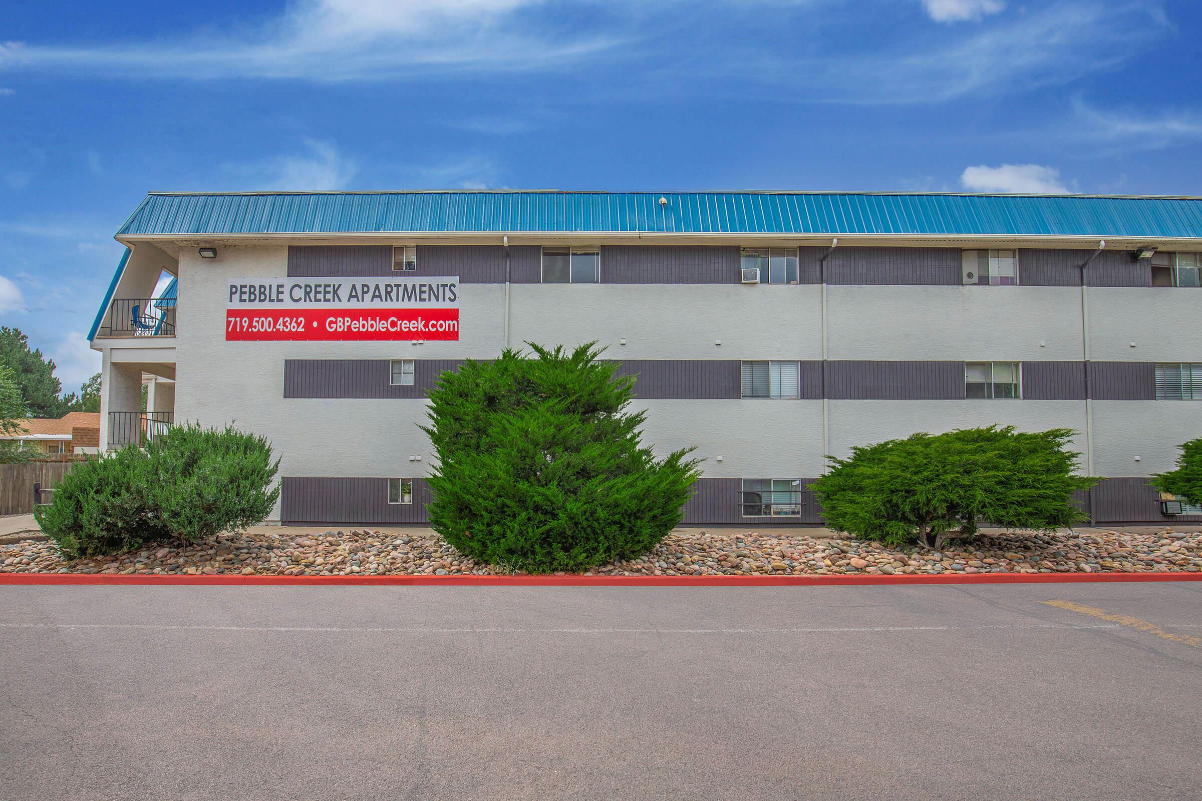 Exterior view of Pebble Creek Apartments, a multi-story building with a teal roof and a sign displaying the name and contact information. Surrounding greenery and a clear blue sky enhance the scene. There is a well-maintained parking area in front.