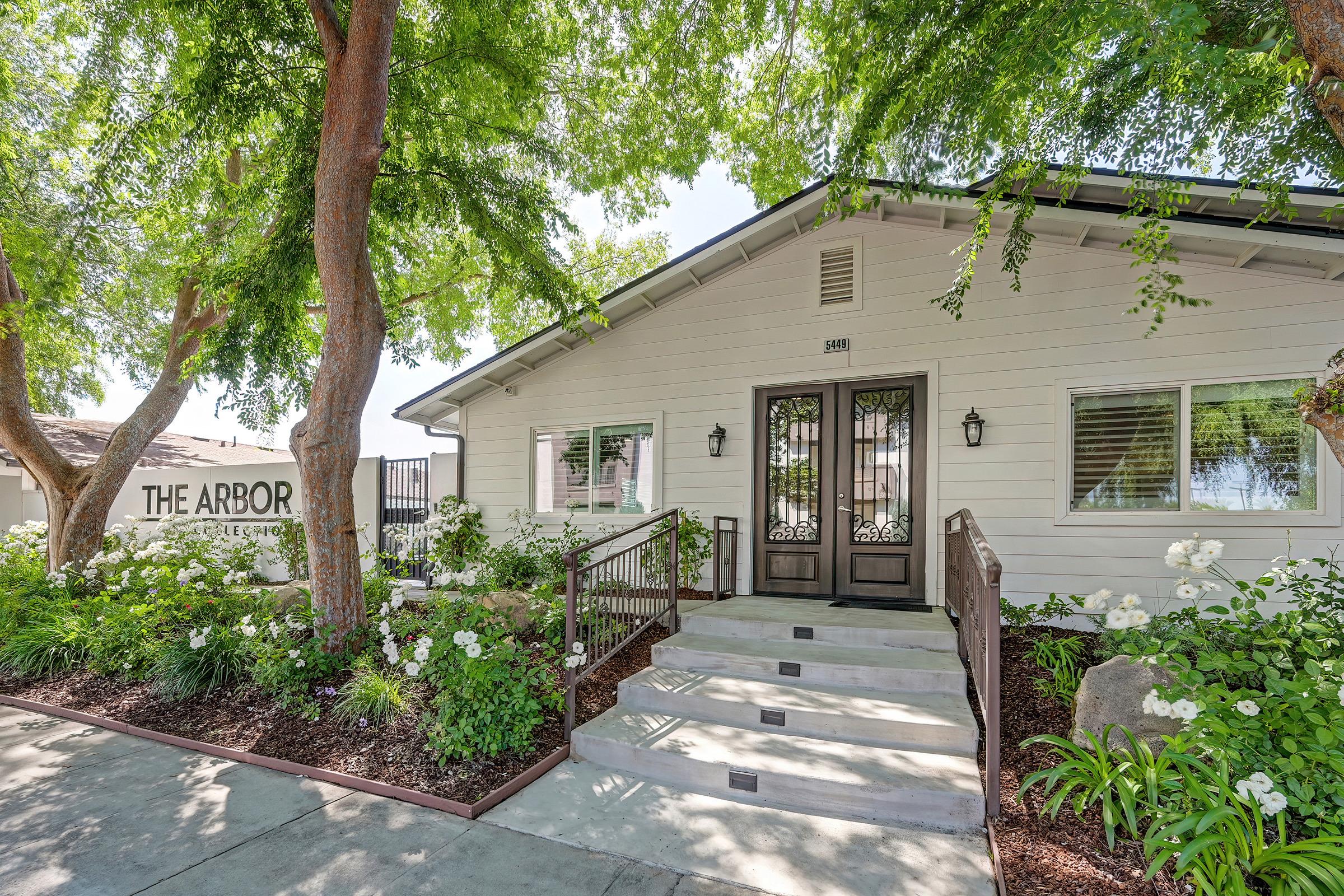 A white building with large front steps and black double doors, surrounded by lush greenery and blooming white flowers. A sign in the background reads "The Arbor." The area is well-maintained, featuring trees that provide shade and a welcoming atmosphere.