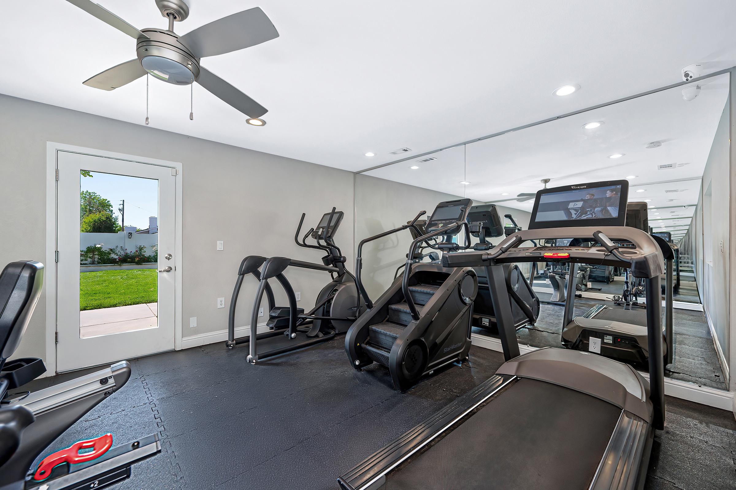 A modern home gym featuring various exercise equipment, including a treadmill and elliptical machine, set against a mirrored wall. A ceiling fan is present, and a door leads to an outdoor view of greenery. The gym has a clean, spacious layout with natural light.