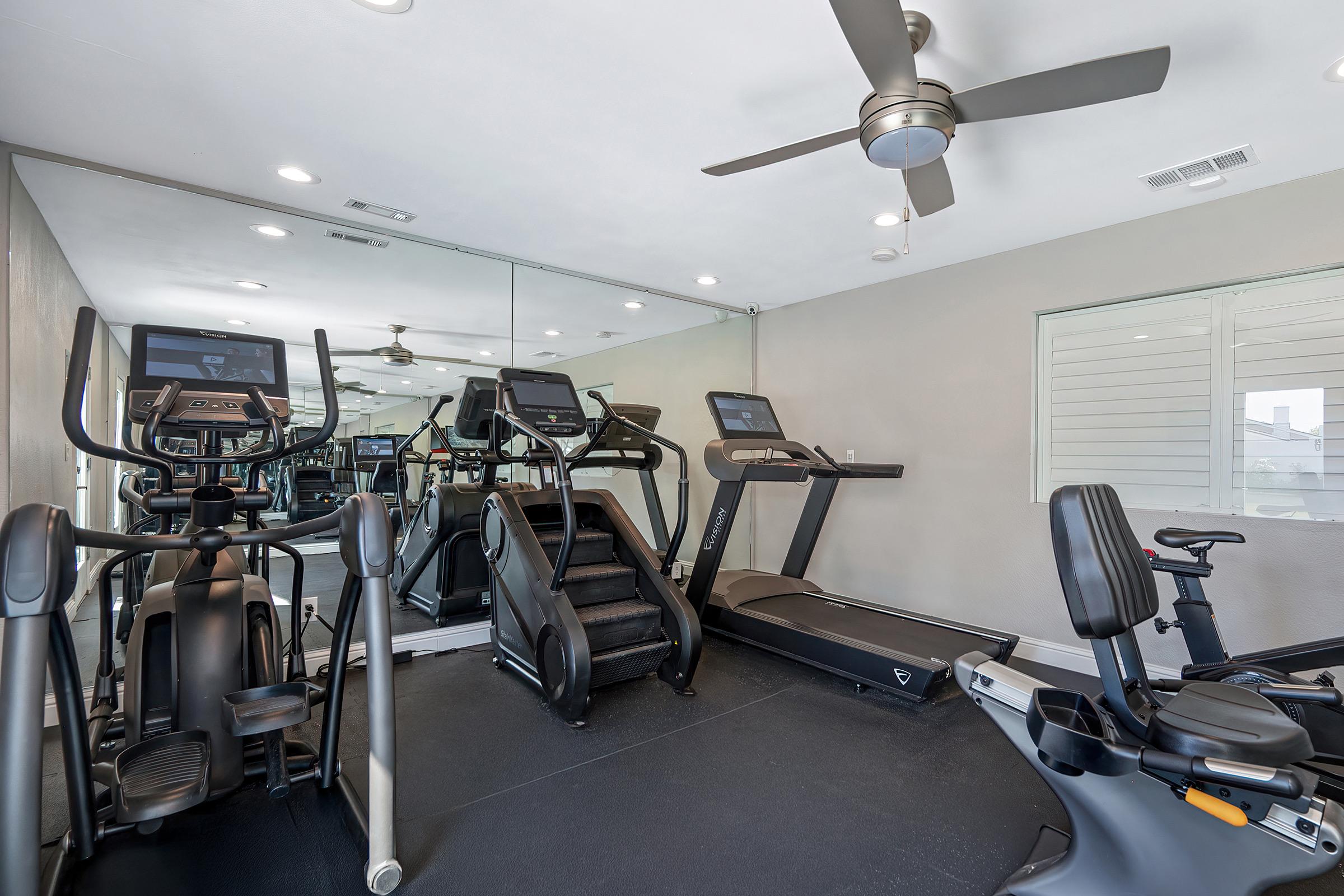 A modern home gym featuring various exercise equipment, including treadmills, an elliptical machine, and a stationary bike. The room has mirrored walls, a ceiling fan, and ample natural light, creating a bright and inviting workout space.