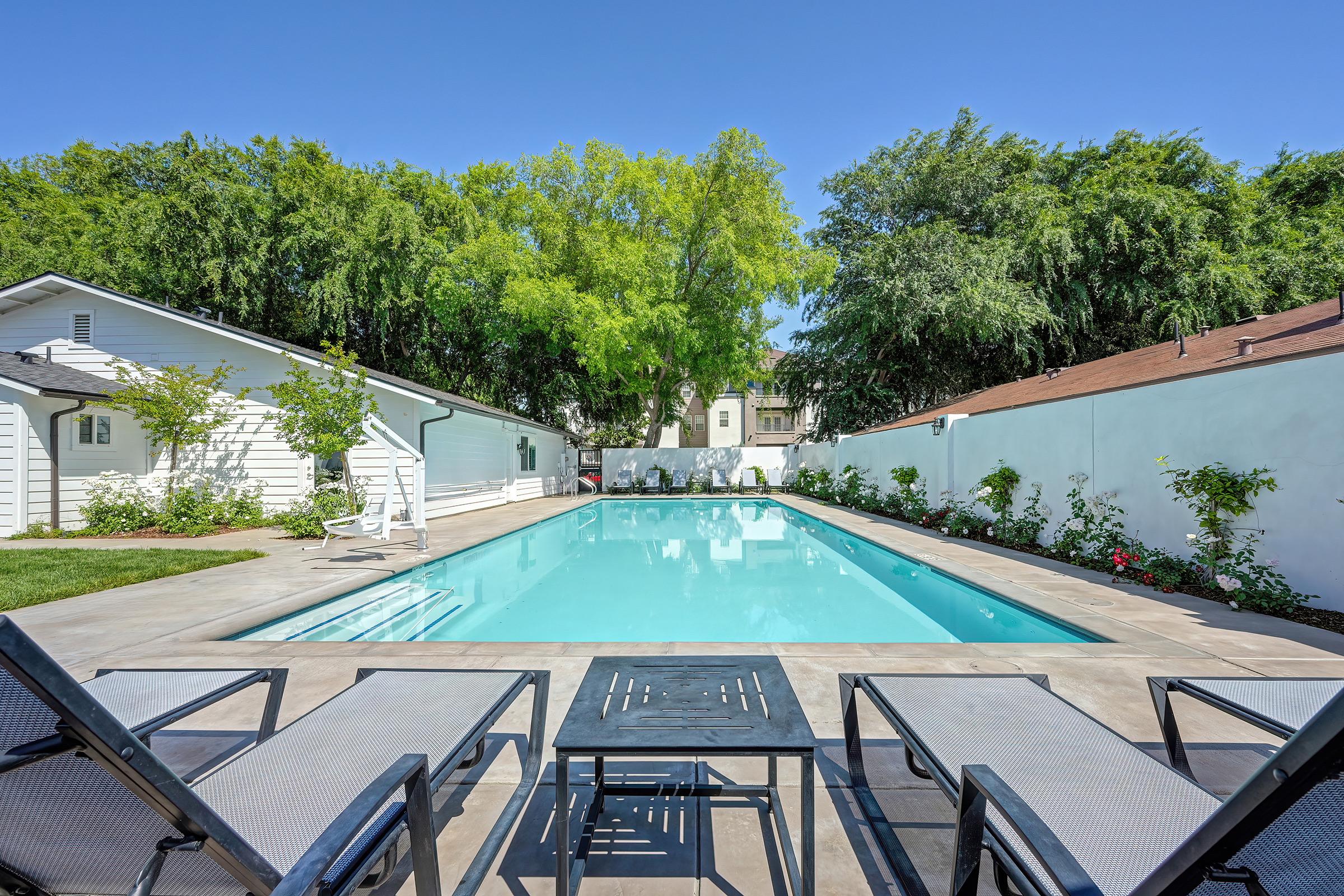 A clear blue swimming pool surrounded by patio furniture, including lounge chairs and a table. Lush green trees and shrubs provide a natural backdrop, with two white buildings flanking the pool area. A sunny sky enhances the serene, inviting atmosphere of this outdoor space.