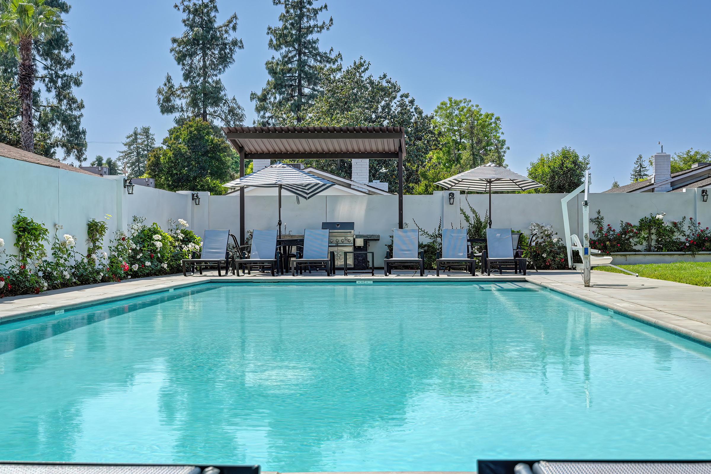A serene backyard scene featuring a crystal-clear swimming pool, surrounded by lounge chairs and shaded by pergolas and umbrellas. Lush greenery with blooming flowers lines the pool area, creating a tranquil and inviting atmosphere under a bright blue sky.
