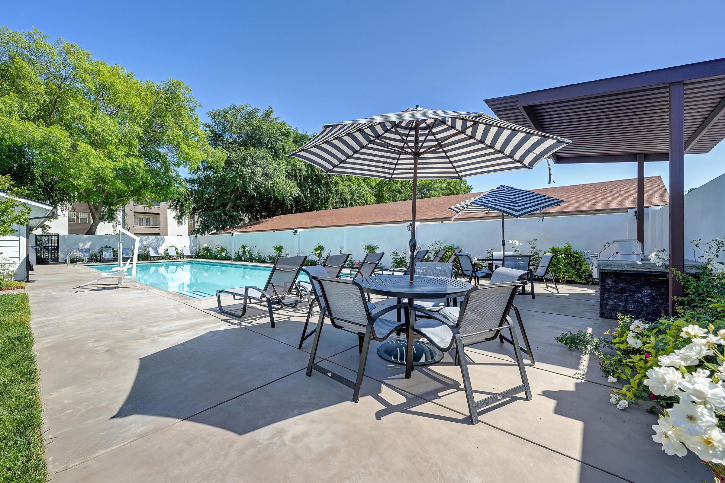 A serene outdoor space featuring a swimming pool surrounded by lounge chairs and tables. Two large umbrellas provide shade, and lush greenery lines the edges, creating a relaxing atmosphere for leisure and gatherings. The clear blue sky adds to the inviting ambiance.