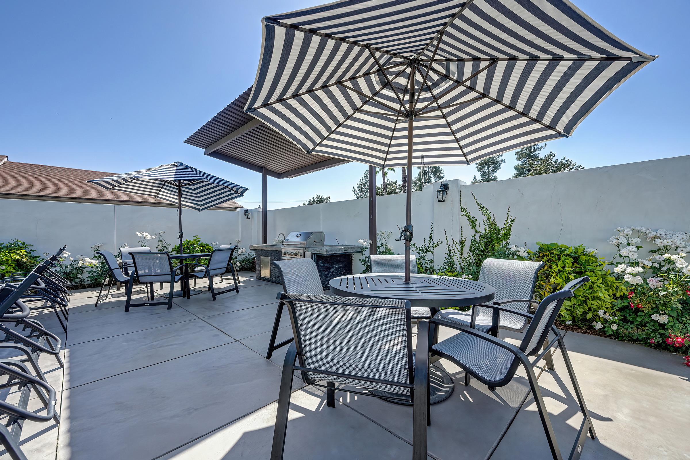 Outdoor patio area featuring several tables and chairs under large striped umbrellas. Lush greenery and flowering plants line the perimeter, with a grill visible in the background. The sky is clear and sunny, creating a bright and inviting atmosphere.