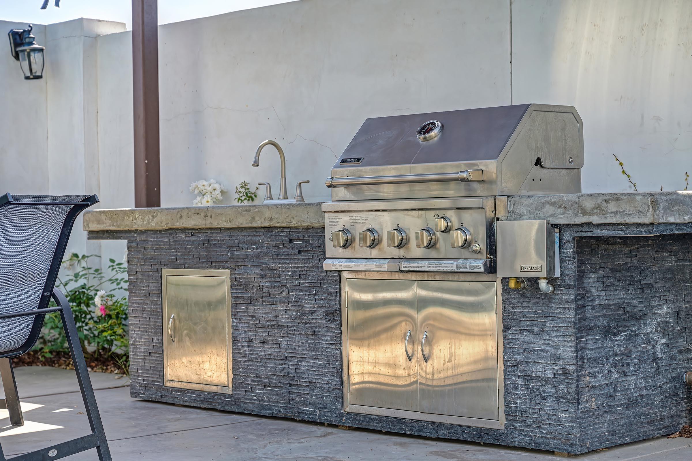 A modern outdoor kitchen featuring a stainless steel gas grill mounted on a stone counter. The grill has multiple burners and knobs for temperature control. Nearby, there is a sink with a faucet, and a stylish chair is positioned in front of the grill, set against a backdrop of a garden.