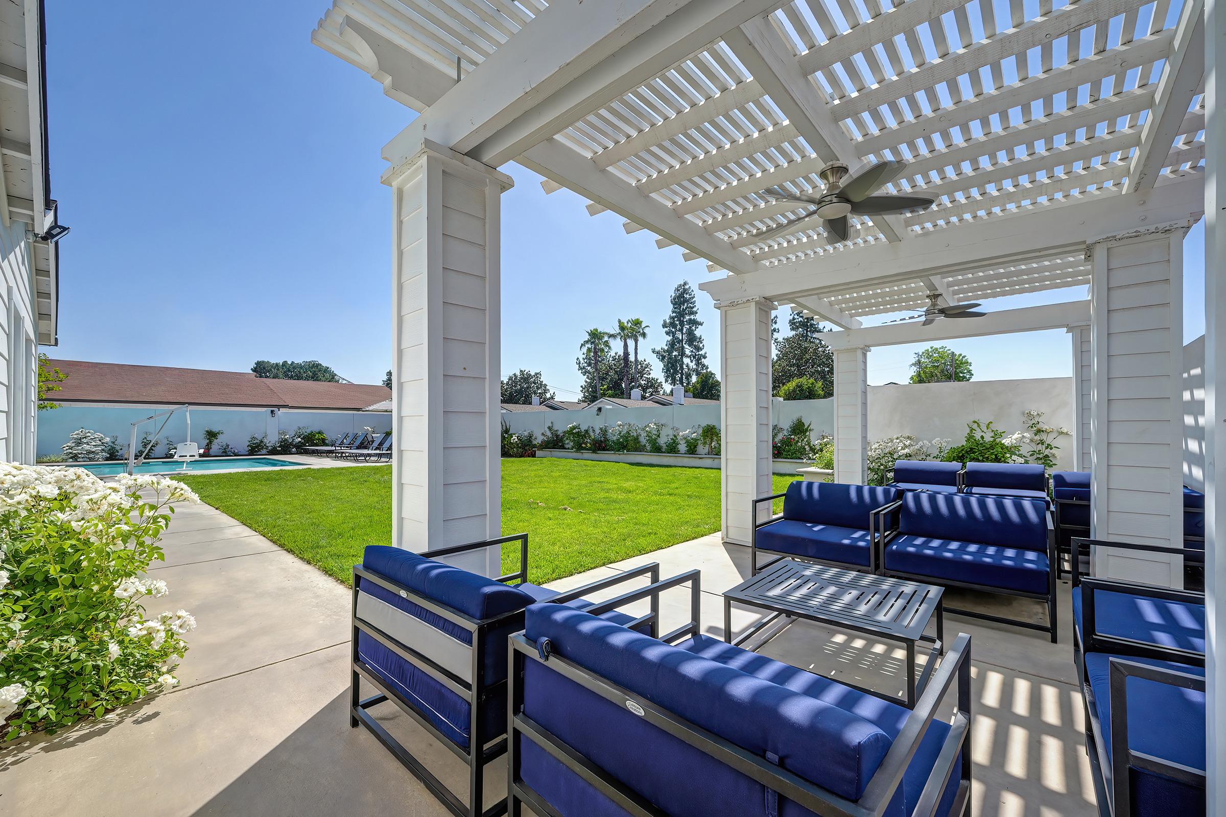 A spacious outdoor patio featuring modern seating with blue cushions, a glass table, and ceiling fans. The area is surrounded by a lush green lawn and flowering bushes. In the background, a swimming pool is visible, creating a relaxing atmosphere for gatherings or leisure activities.