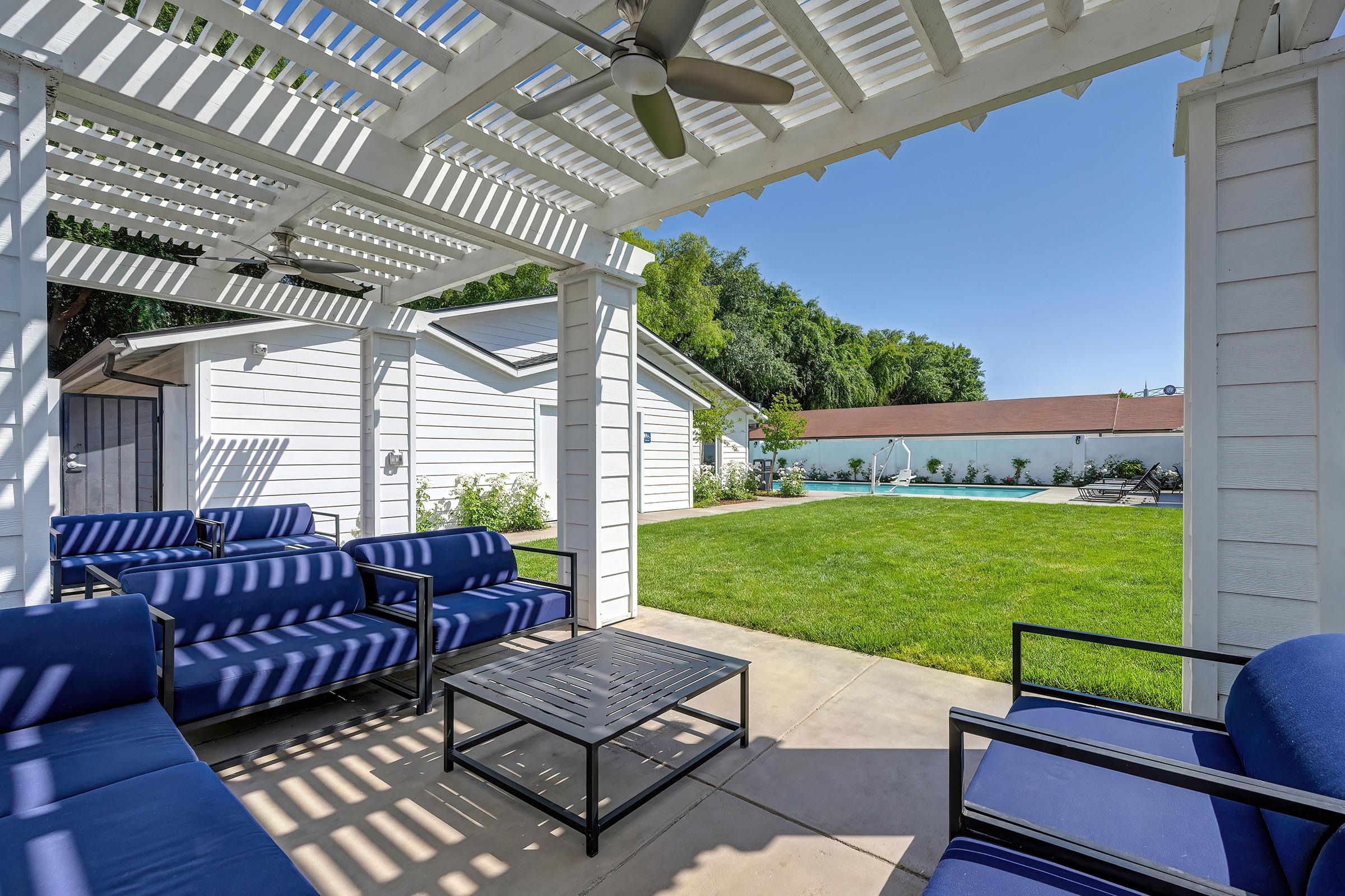 A shaded outdoor patio area with blue furniture, including chairs and a table, overlooking a green lawn and a swimming pool in the background. The structure features a white wooden pergola and well-maintained landscaping, creating a serene and inviting space for relaxation.