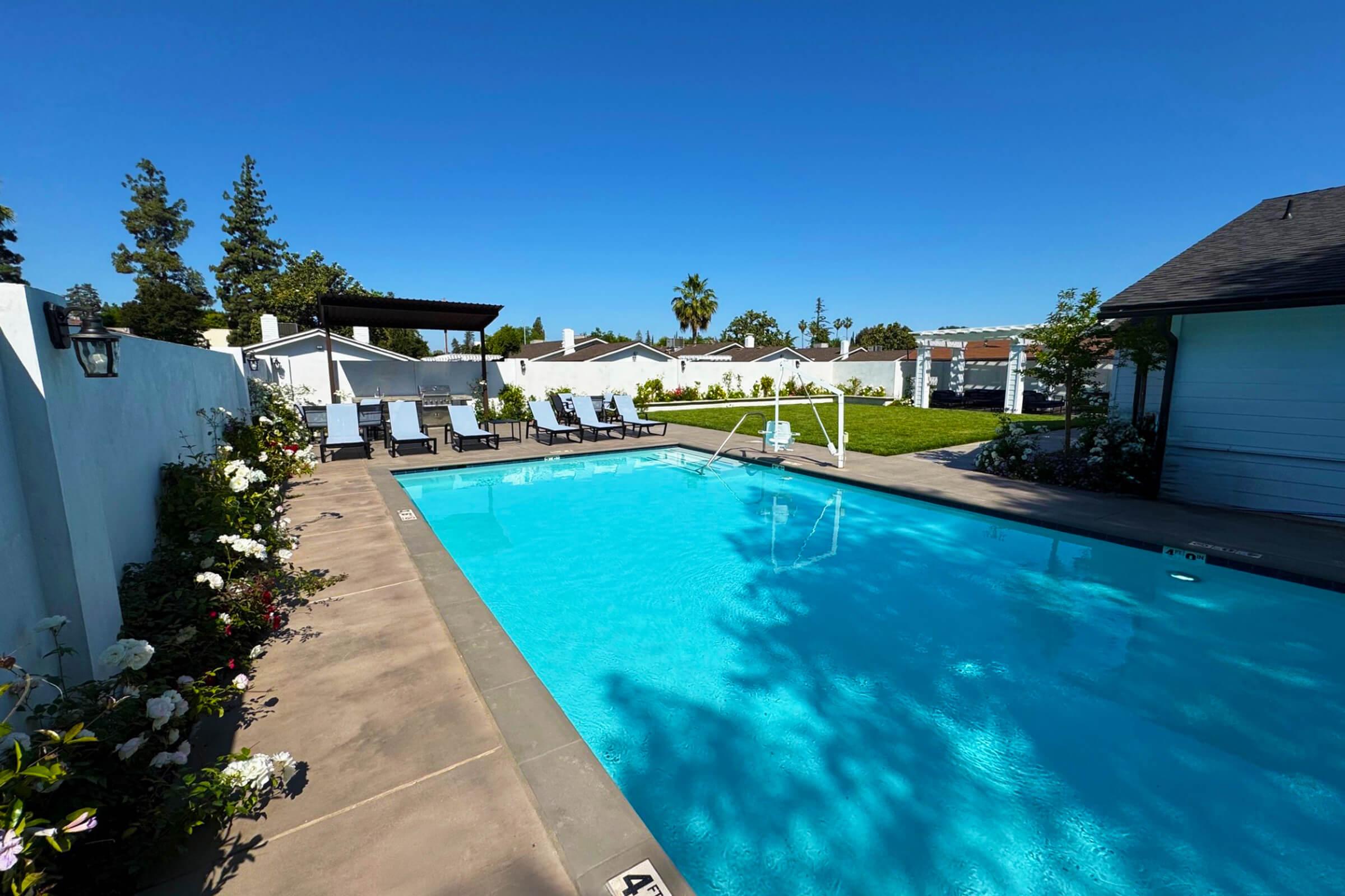A bright, inviting swimming pool surrounded by lounge chairs and a well-maintained garden. The clear blue water reflects the sunny sky, with trees and houses in the background. A shaded pergola provides a relaxing spot nearby, creating a tranquil outdoor space.